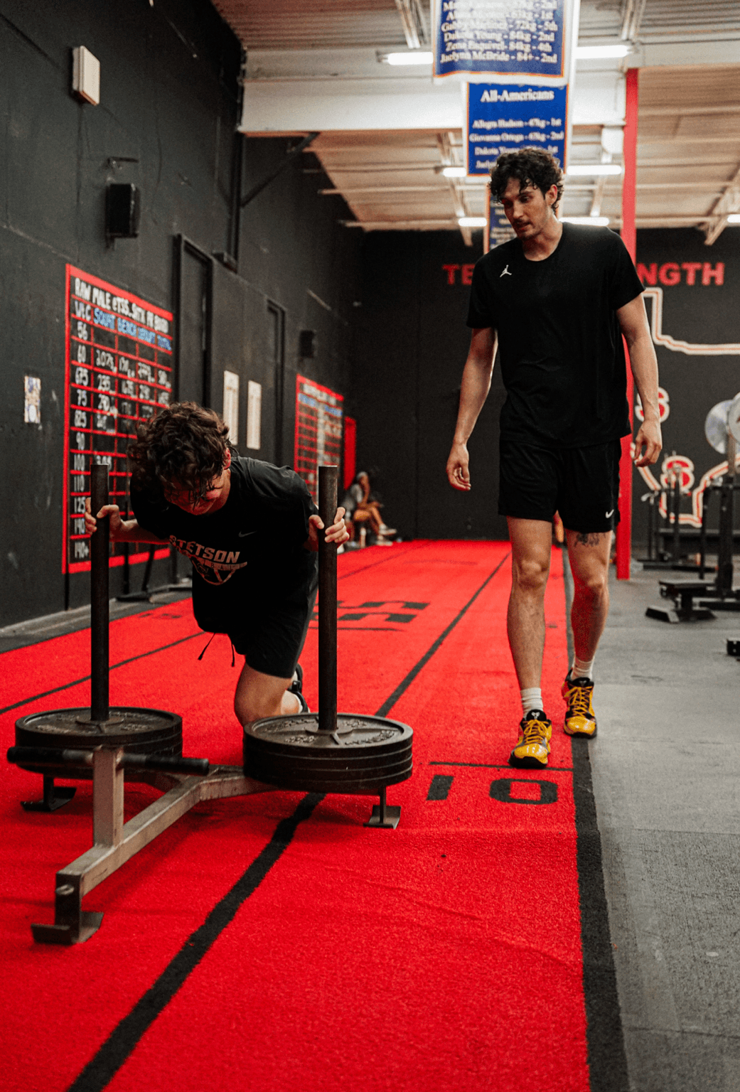 Two men in a gym, one pushing weighted sled, the other walking beside him on a red track, gym equipment in background.