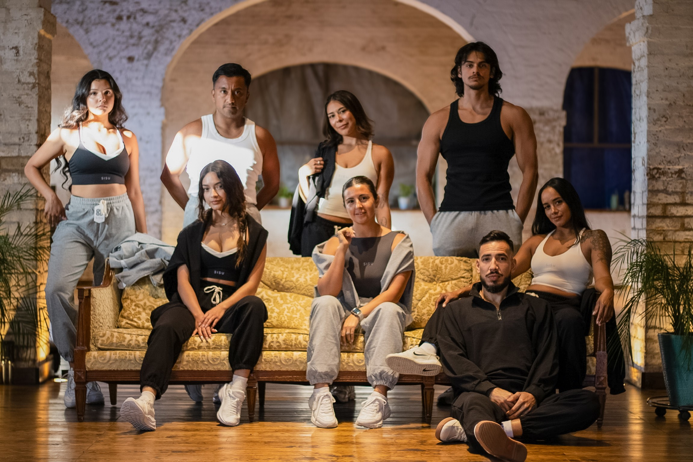 Group of eight people posing on a stage with a stone arch background, some sitting and some standing, in casual clothing.