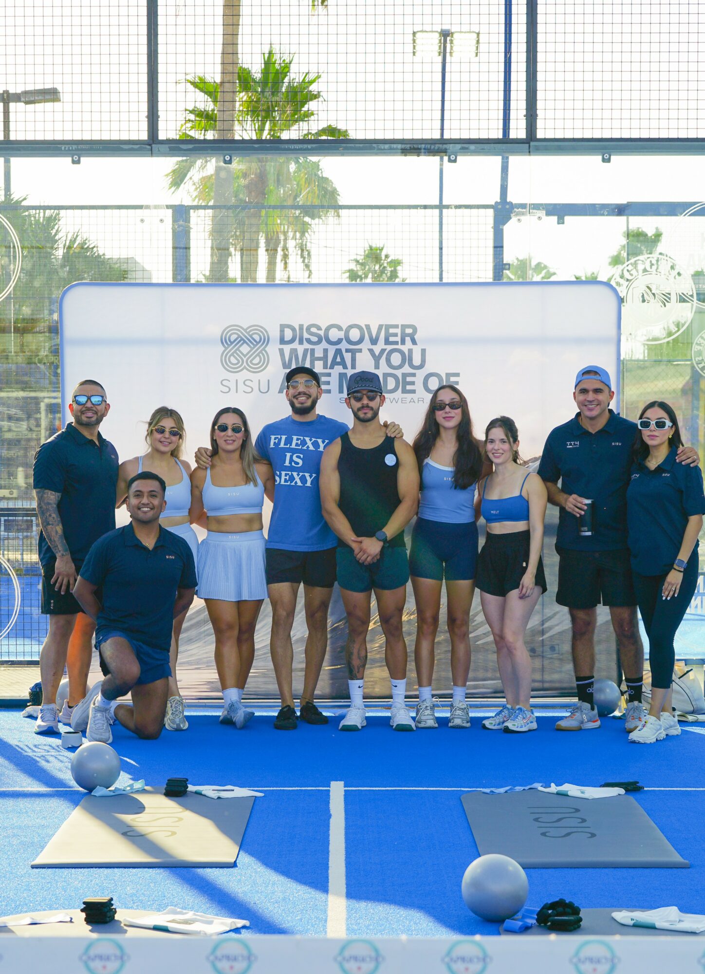 Group of eight people standing on a blue indoor sports court, some wearing sunglasses and athletic clothing, with a large window and palm trees in the background.