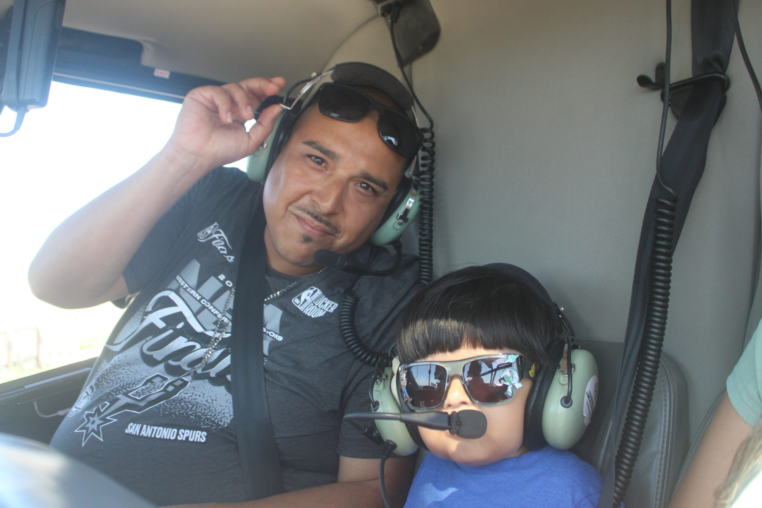 Man and child wearing headsets sit inside a vehicle, with the man adjusting his headset and both looking at the camera.