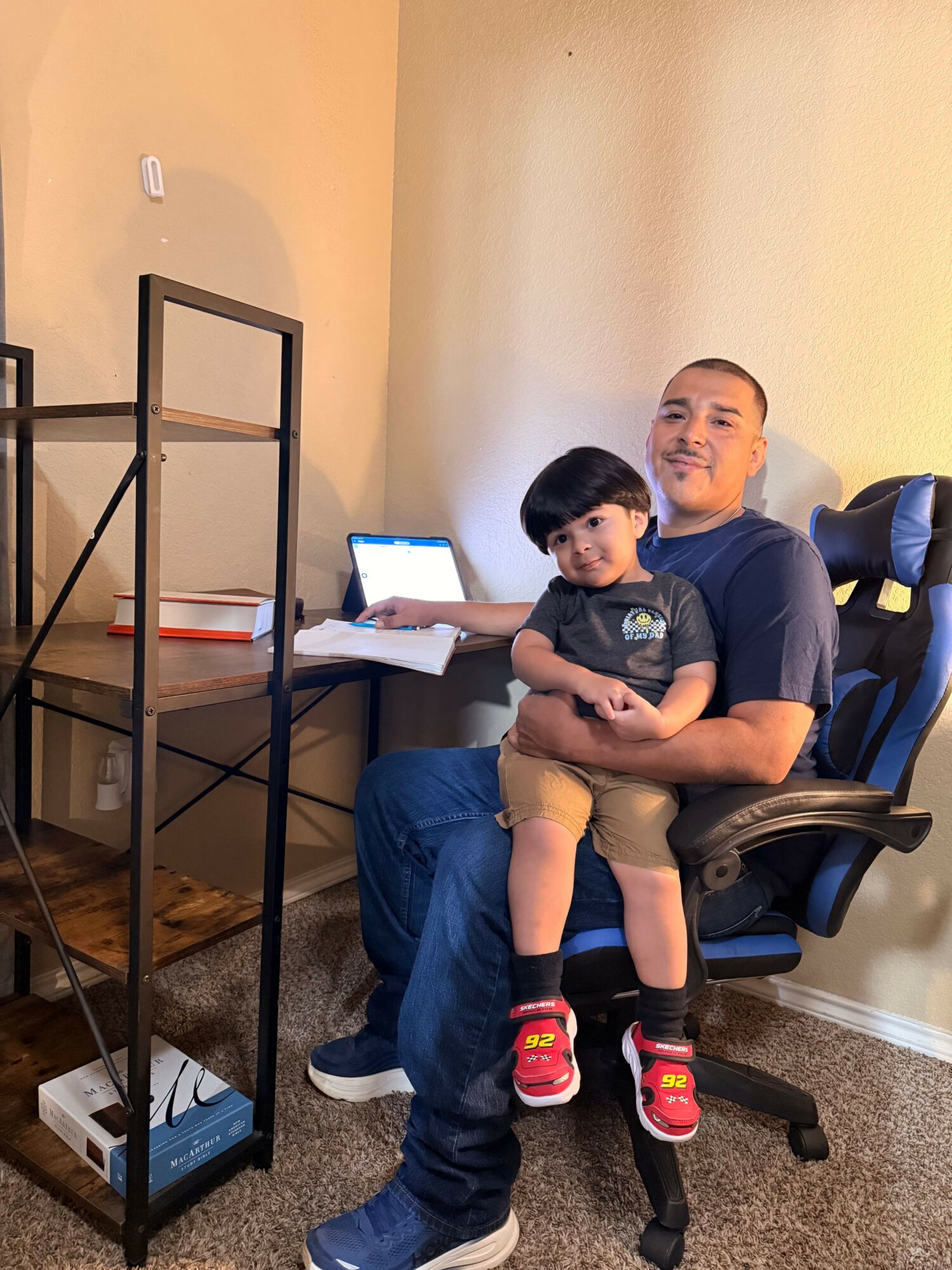 Two people, one adult and one child, sitting together in a room with a computer and a glass display case.