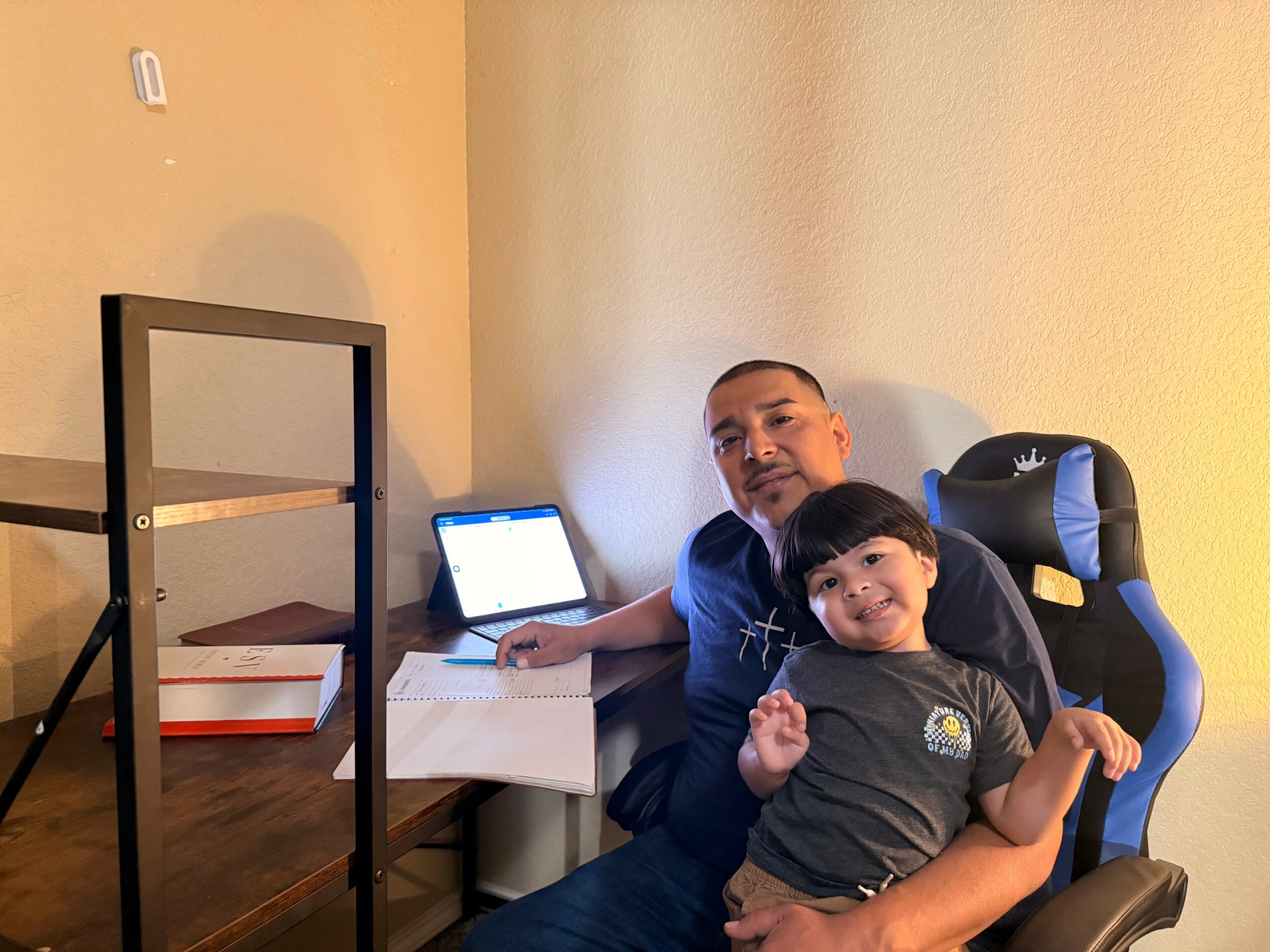 Man and child sitting together in a room, with a laptop on a small table nearby, smiling at the camera.