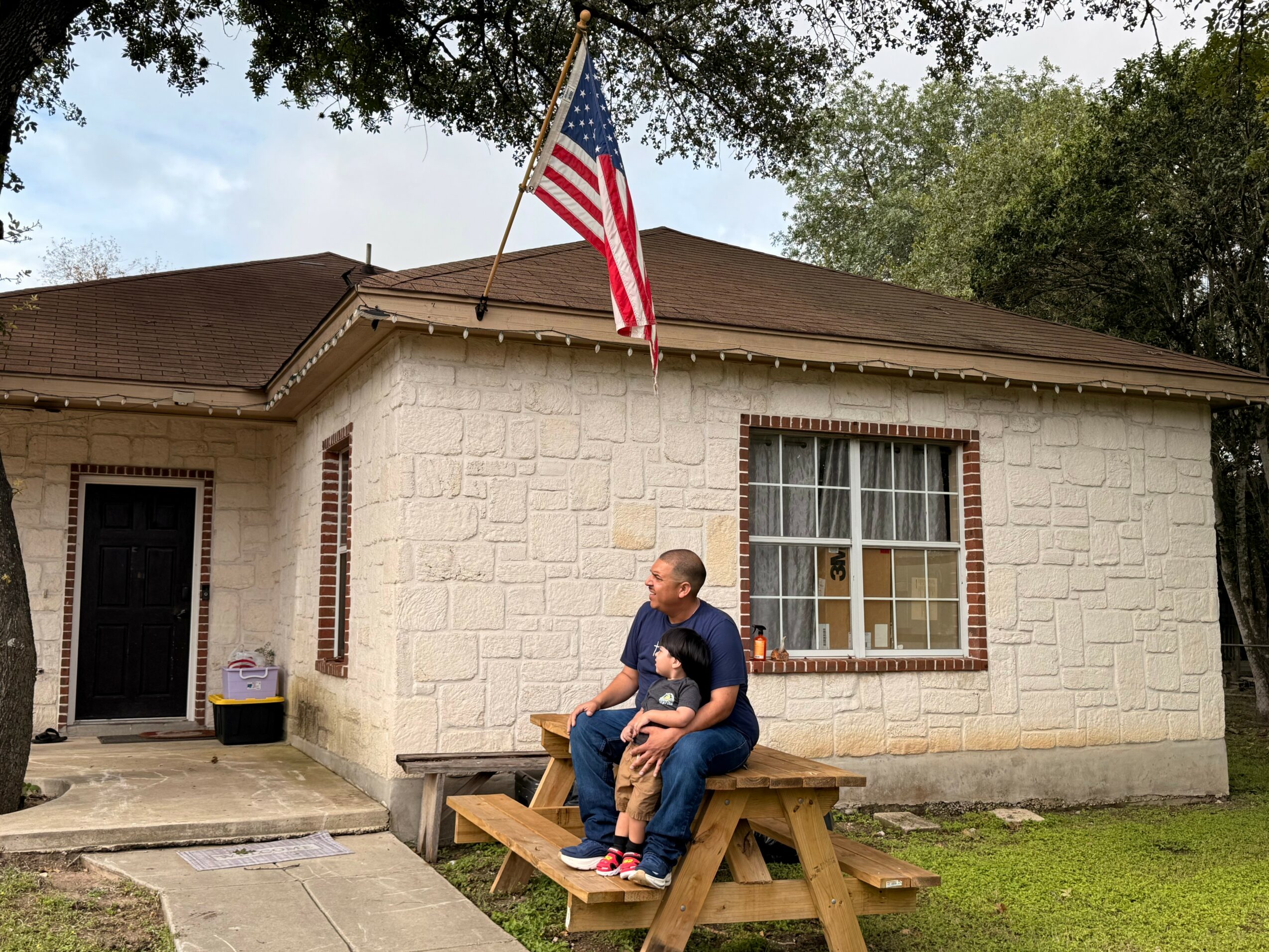 Person sitting on a picnic table in front of a house with an American flag, window, and door, surrounded by trees.