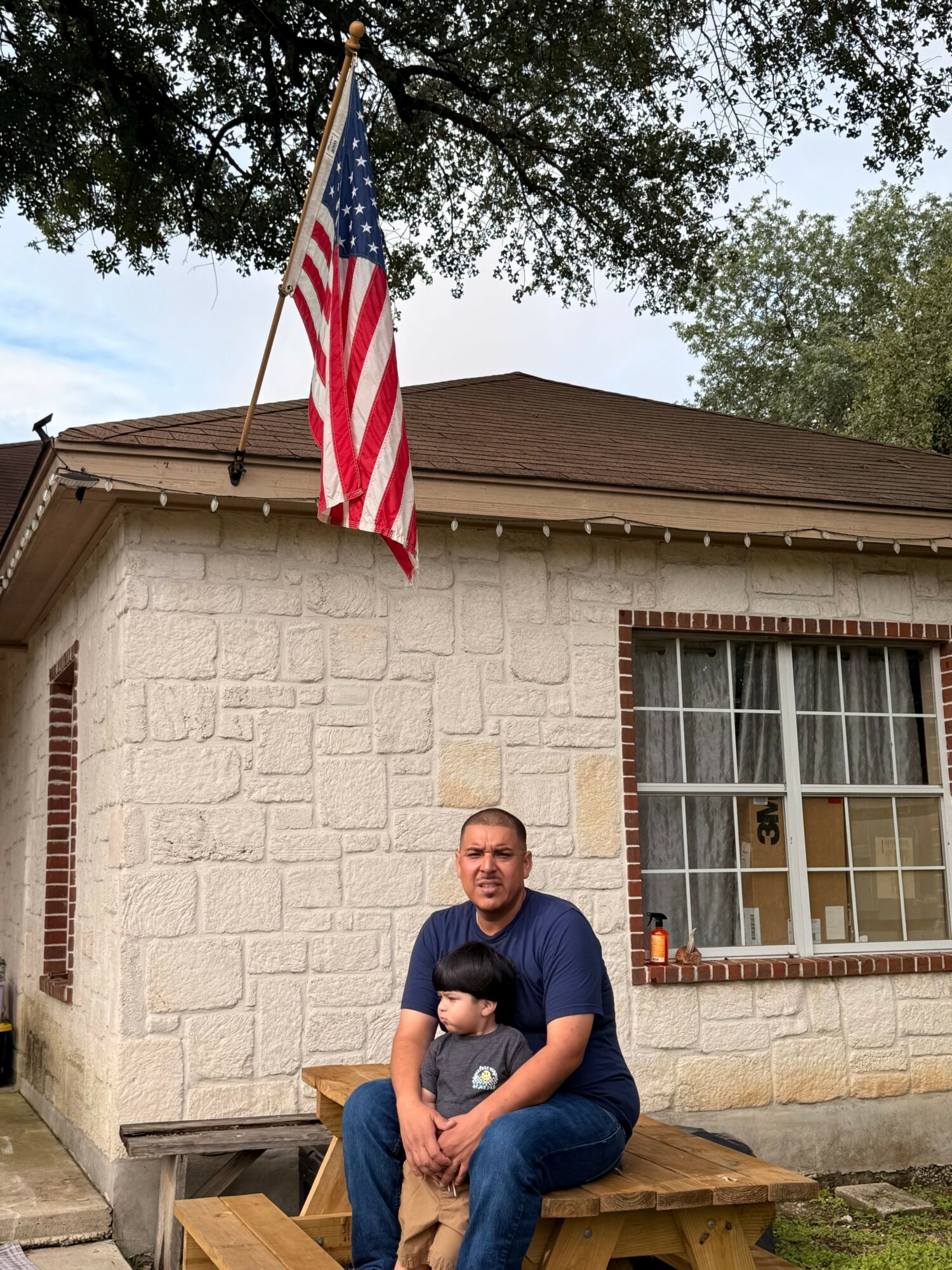 Person sitting on a bench outside a building with an American flag, trees, and a window behind them.