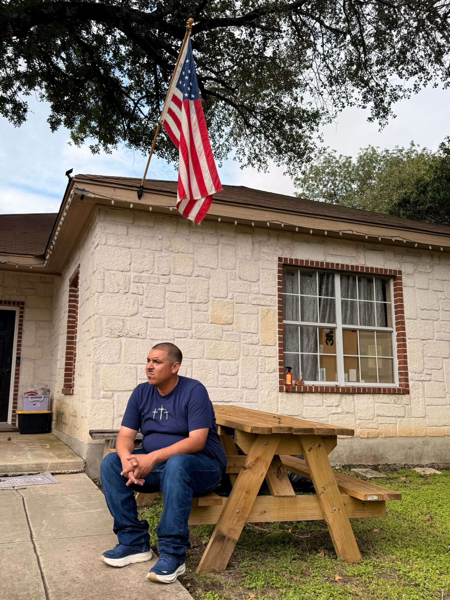 Man sitting on a wooden bench outside a brick building with an American flag, window, and green grass.
