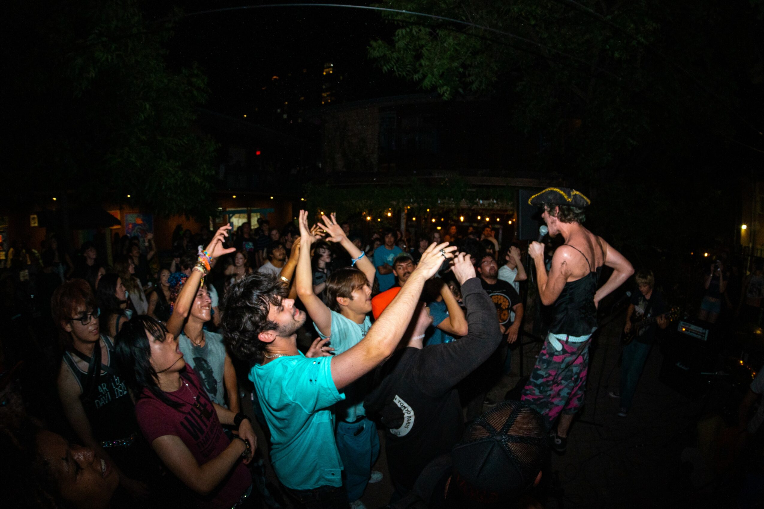 People dancing outdoors at night, some with hands raised, in a lively crowd with trees and lights in the background.