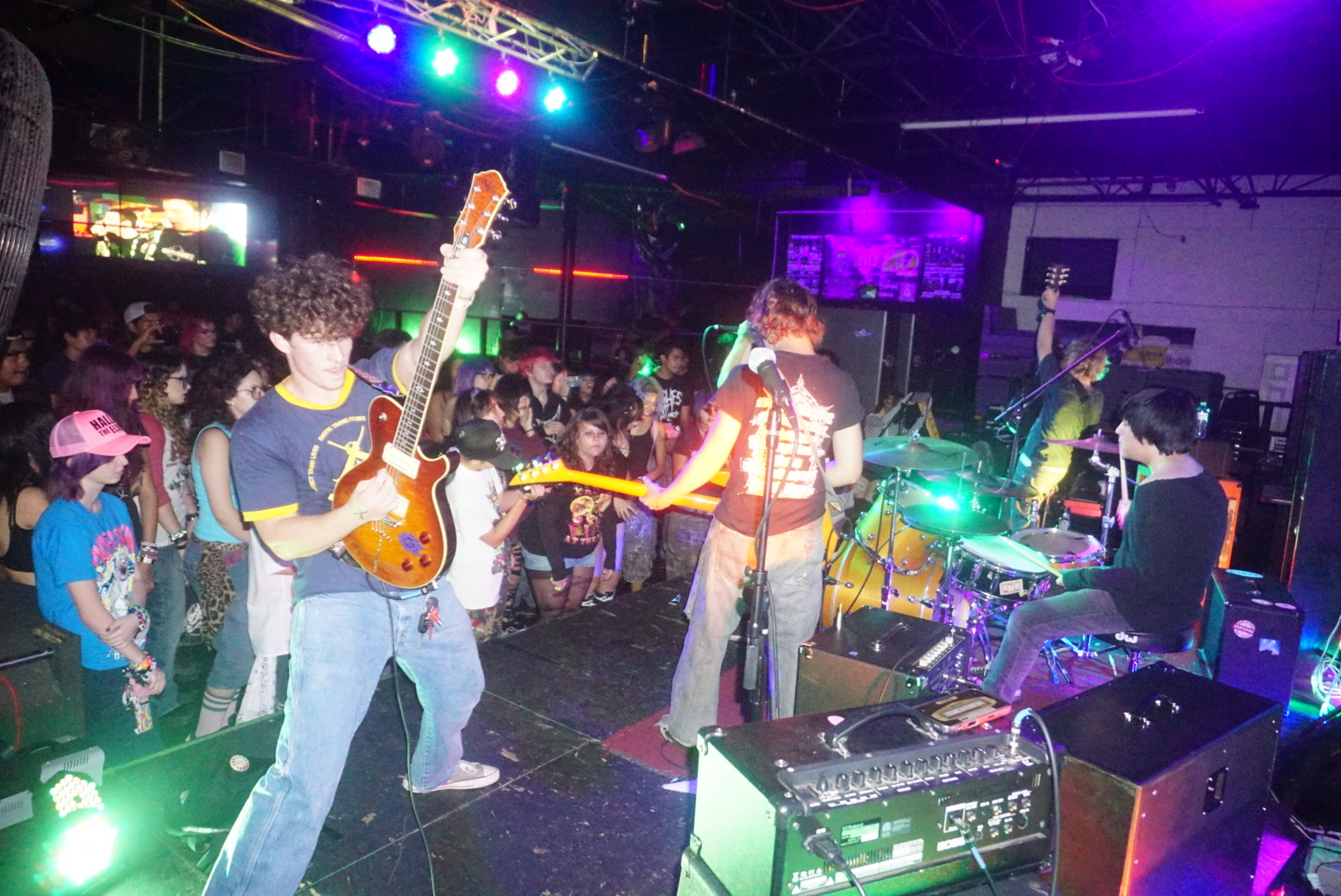Band performs on stage with audience watching, colorful lights illuminate the scene, musical equipment visible, in a dark indoor venue.