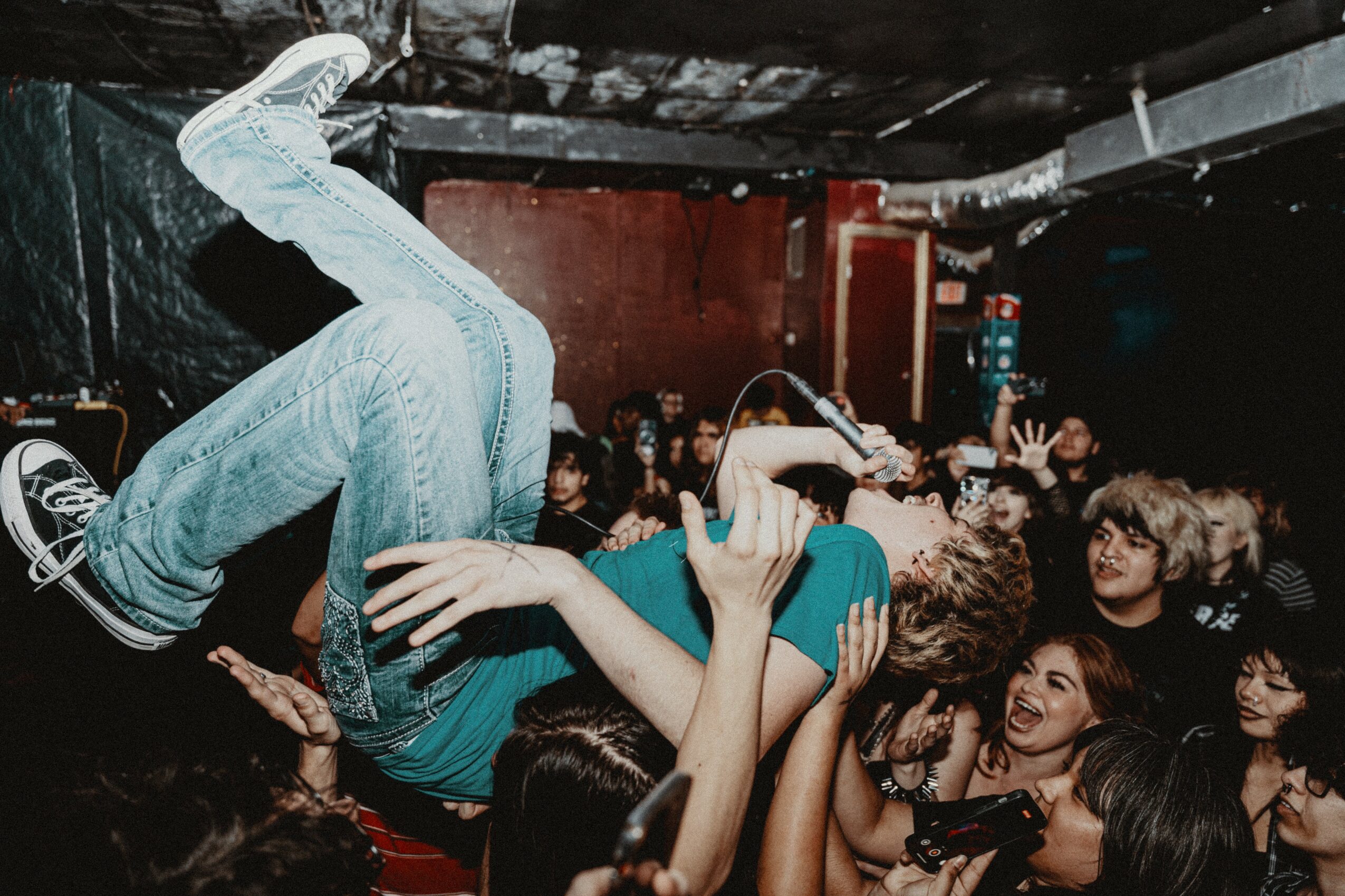 Person performing a flip above a crowd at a concert or event, with audience members reaching up and smiling.