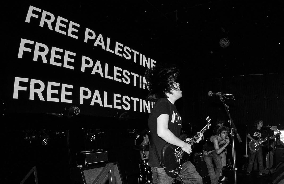 Musicians perform on stage with a large screen displaying 'FREE PALESTINE' in the background.