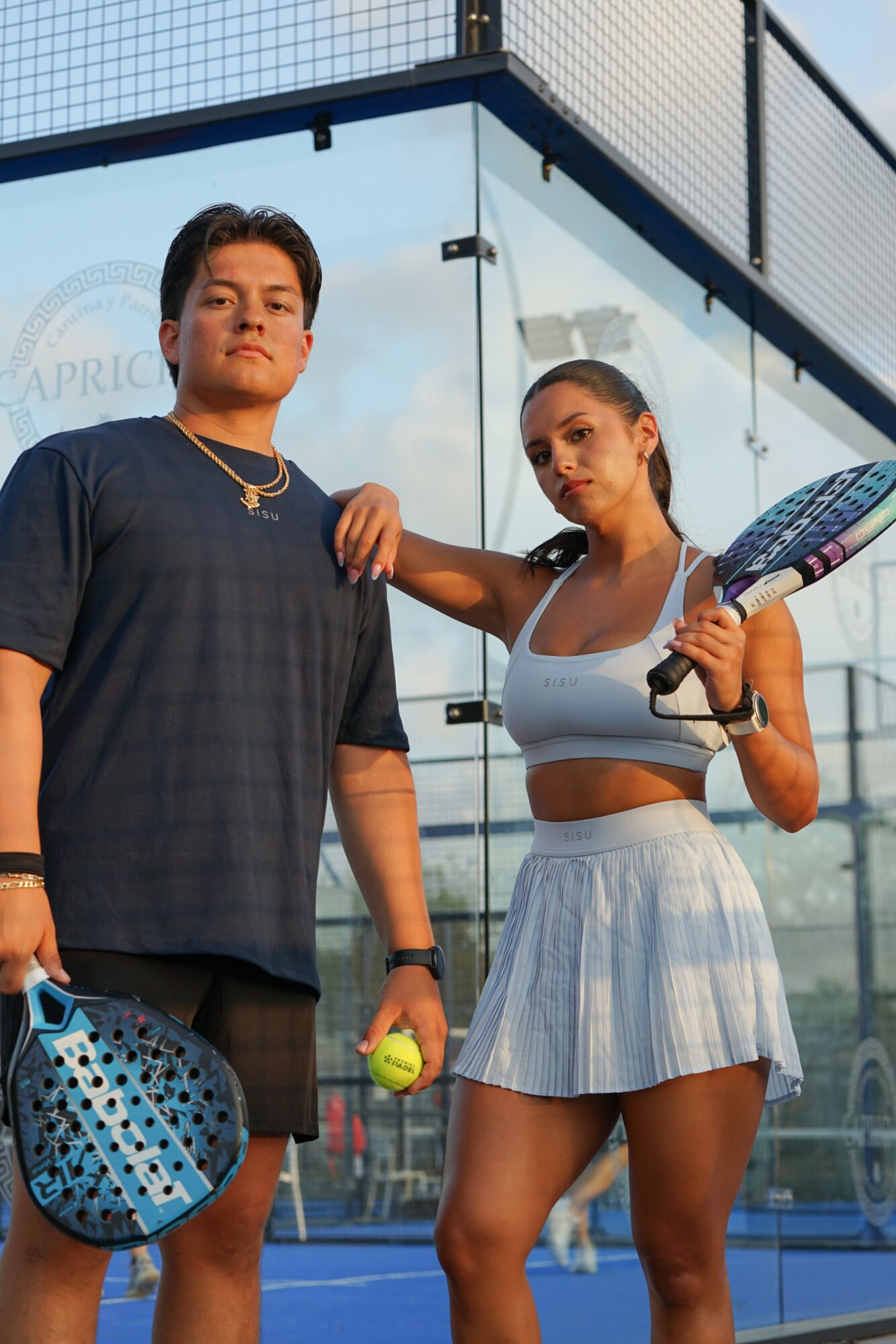 Two young women on a tennis court, one holding a tennis racket and ball, the other with a tennis racket and helmet, standing outdoors.
