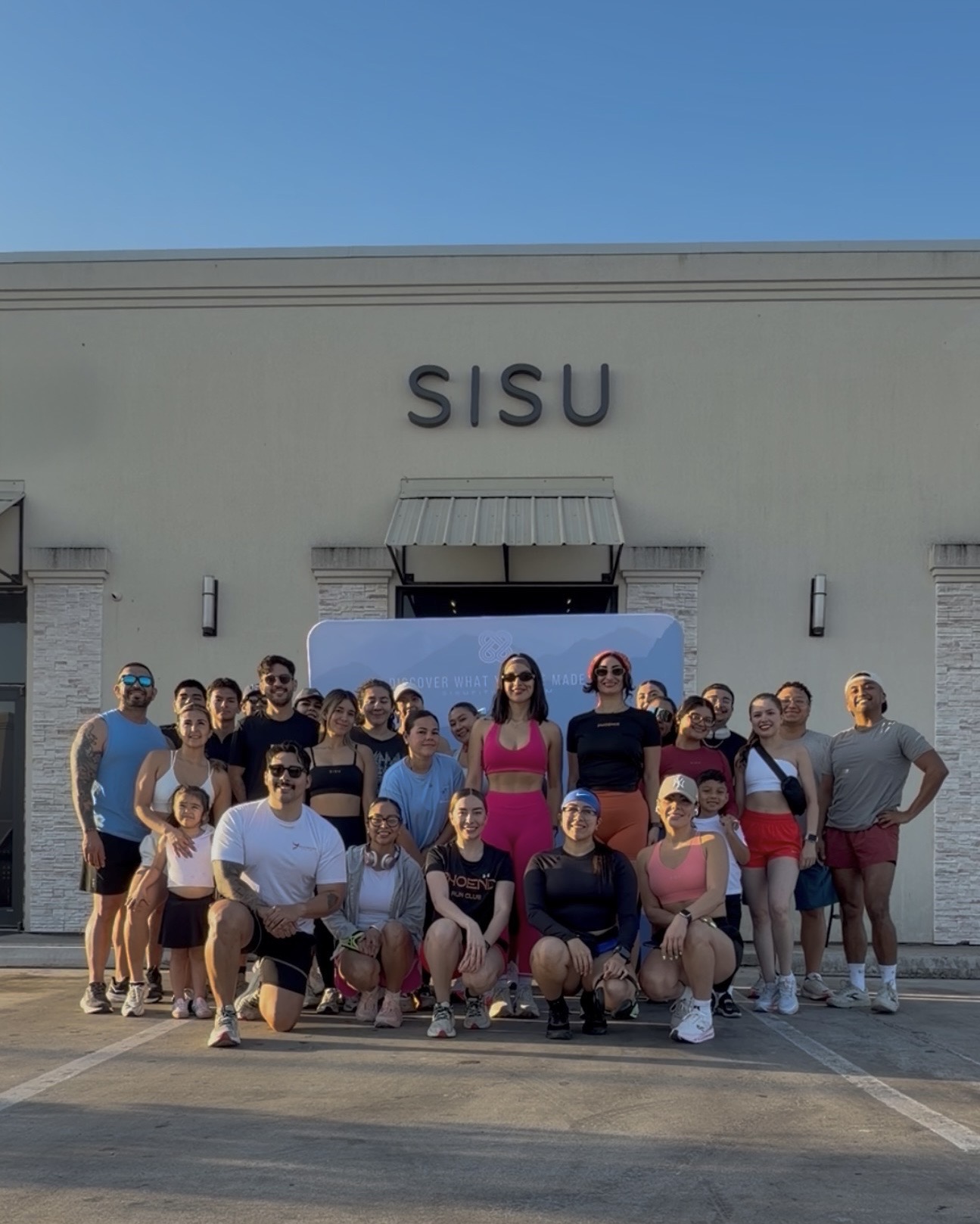 Group of people standing and sitting in front of a building with 'SISU' sign, under a clear blue sky.