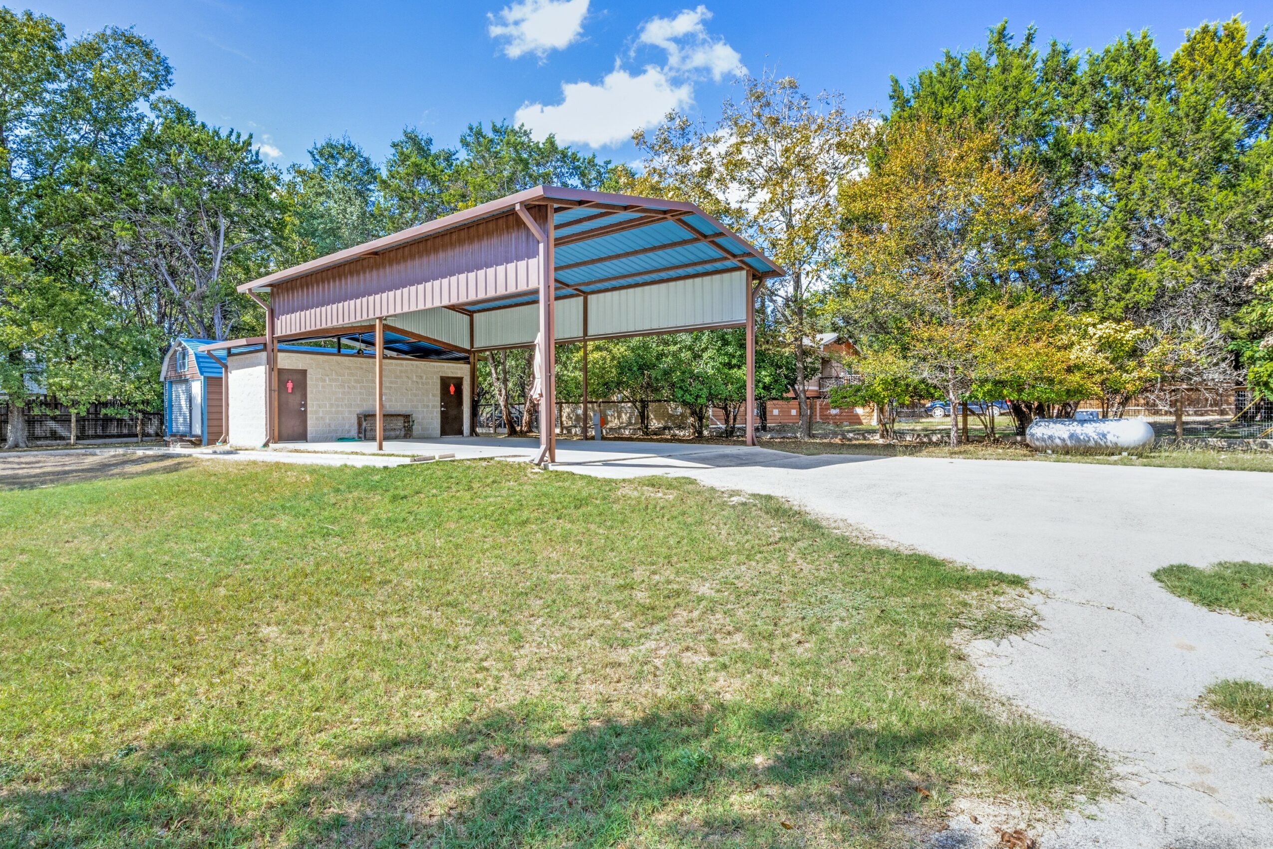 Open-sided shelter with a metal roof and supporting columns, surrounded by grass and trees, under a blue sky with clouds.