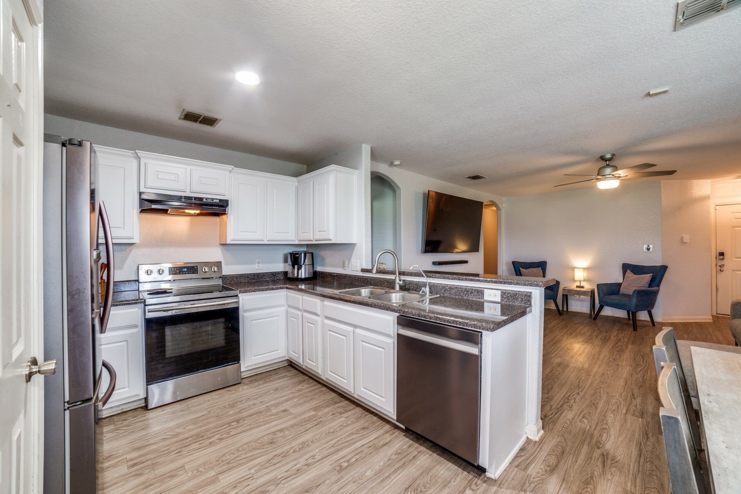 Kitchen with white cabinets, stainless steel appliances, and a granite countertop island, open to a living area with chairs and a TV.