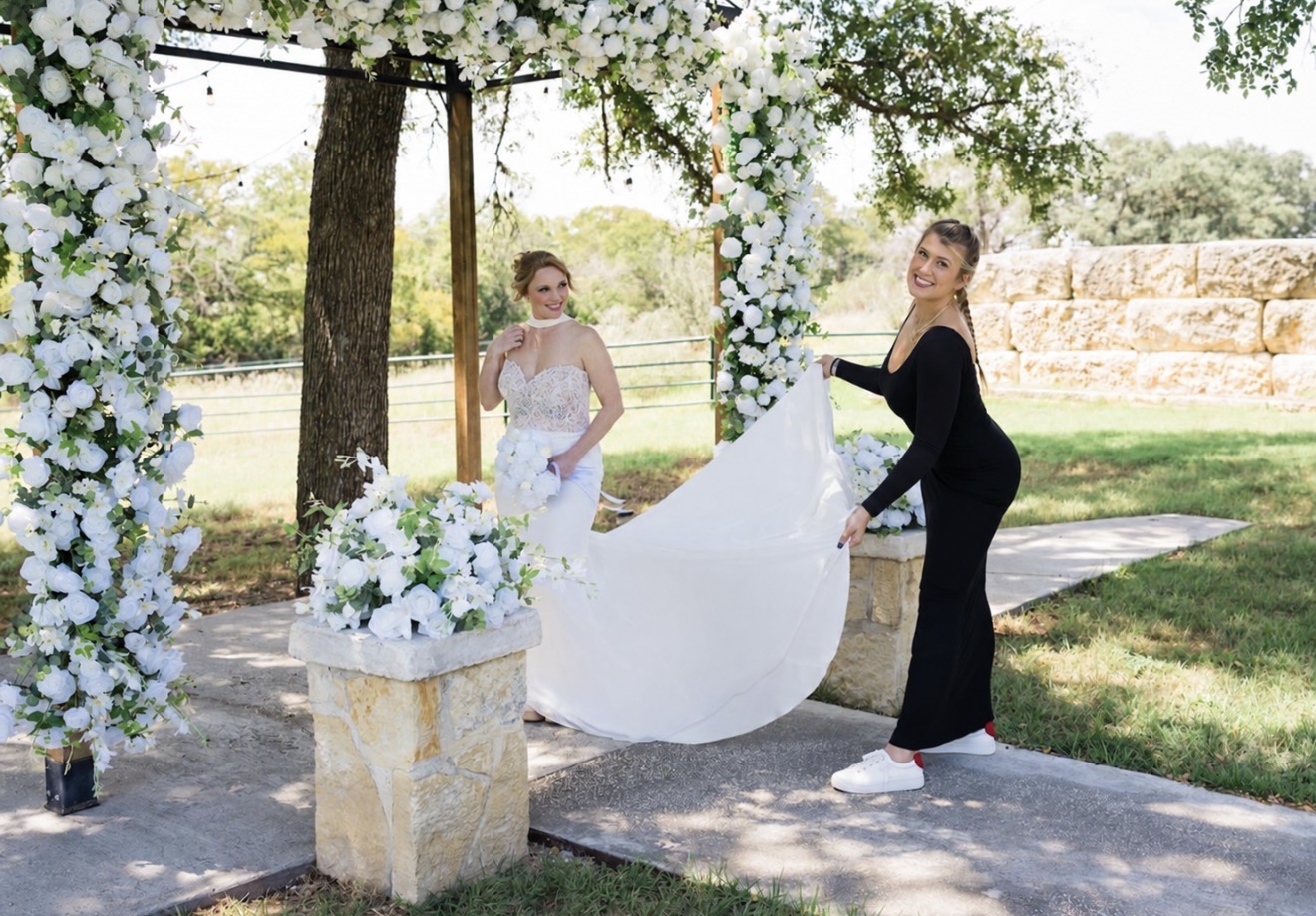 Two women outdoors near a floral arch, one holding a white fabric, with trees and a stone wall in background.
