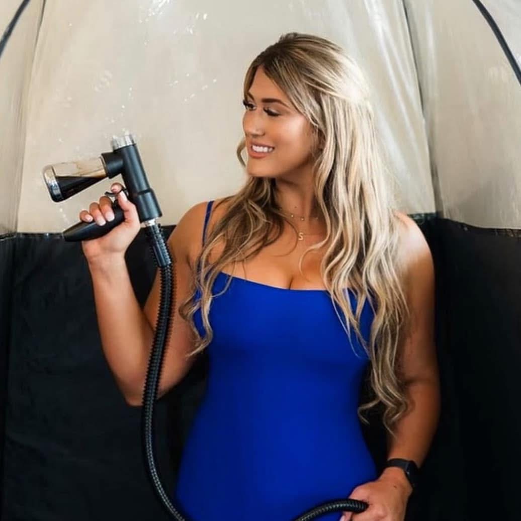Young woman with long blonde hair smiling, holding a showerhead, wearing a blue dress inside a tent.