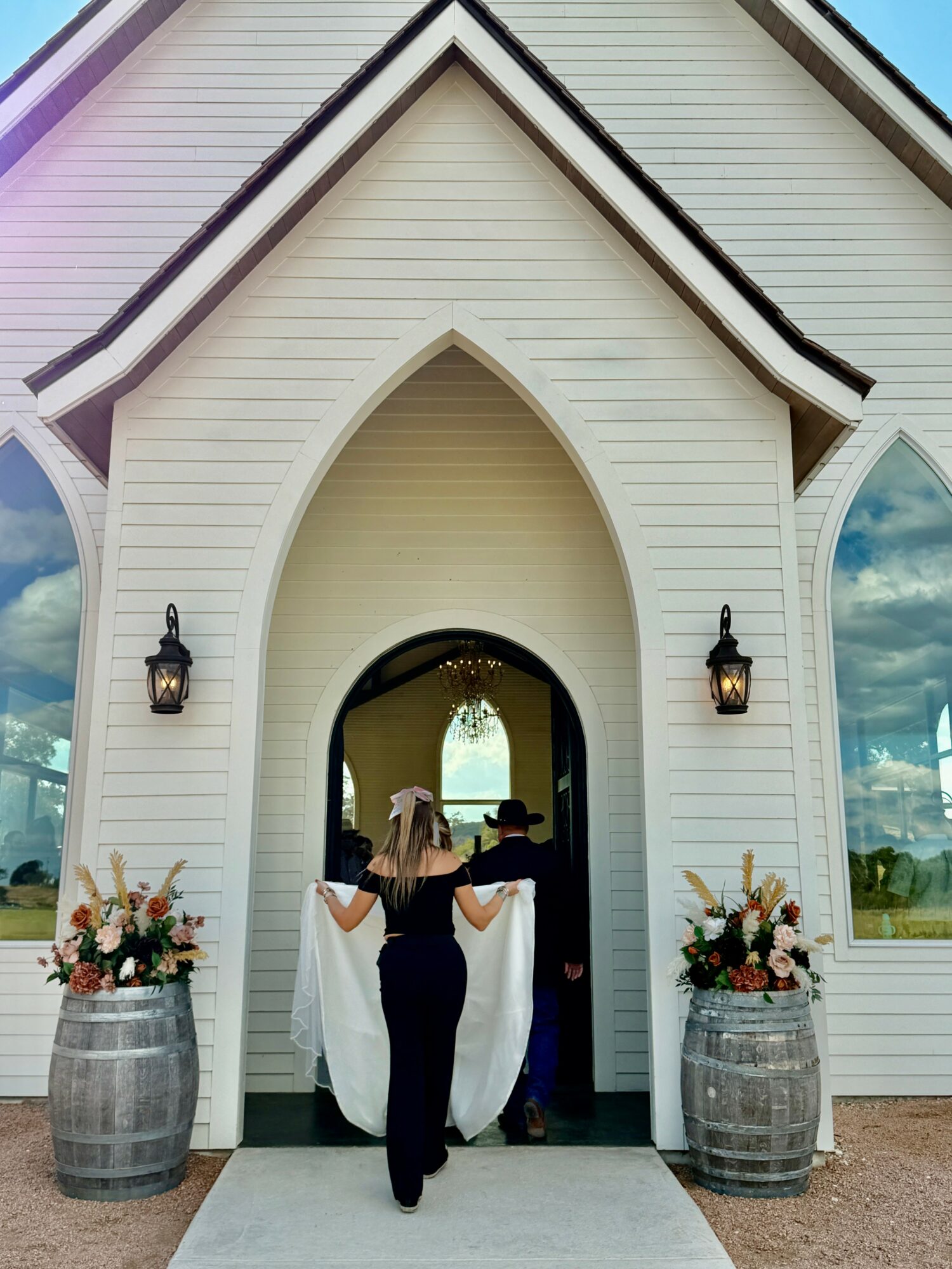 Person standing at church entrance with floral arrangements in barrels, two lanterns on sides, arched doorway, and reflection in windows