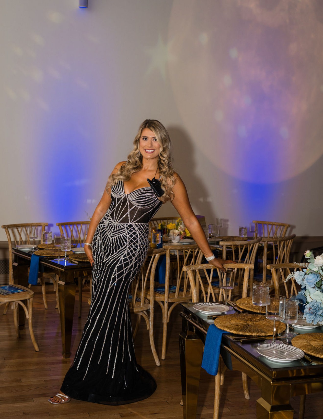 Woman in a black and white patterned gown standing in a decorated event space with tables and chairs, smiling.