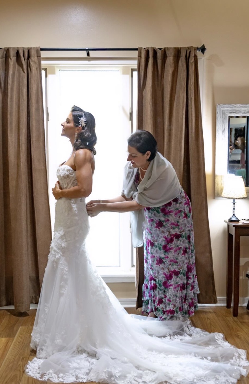 Two women in a room, one in a wedding dress, the other helping her with the dress, standing near a door with curtains.