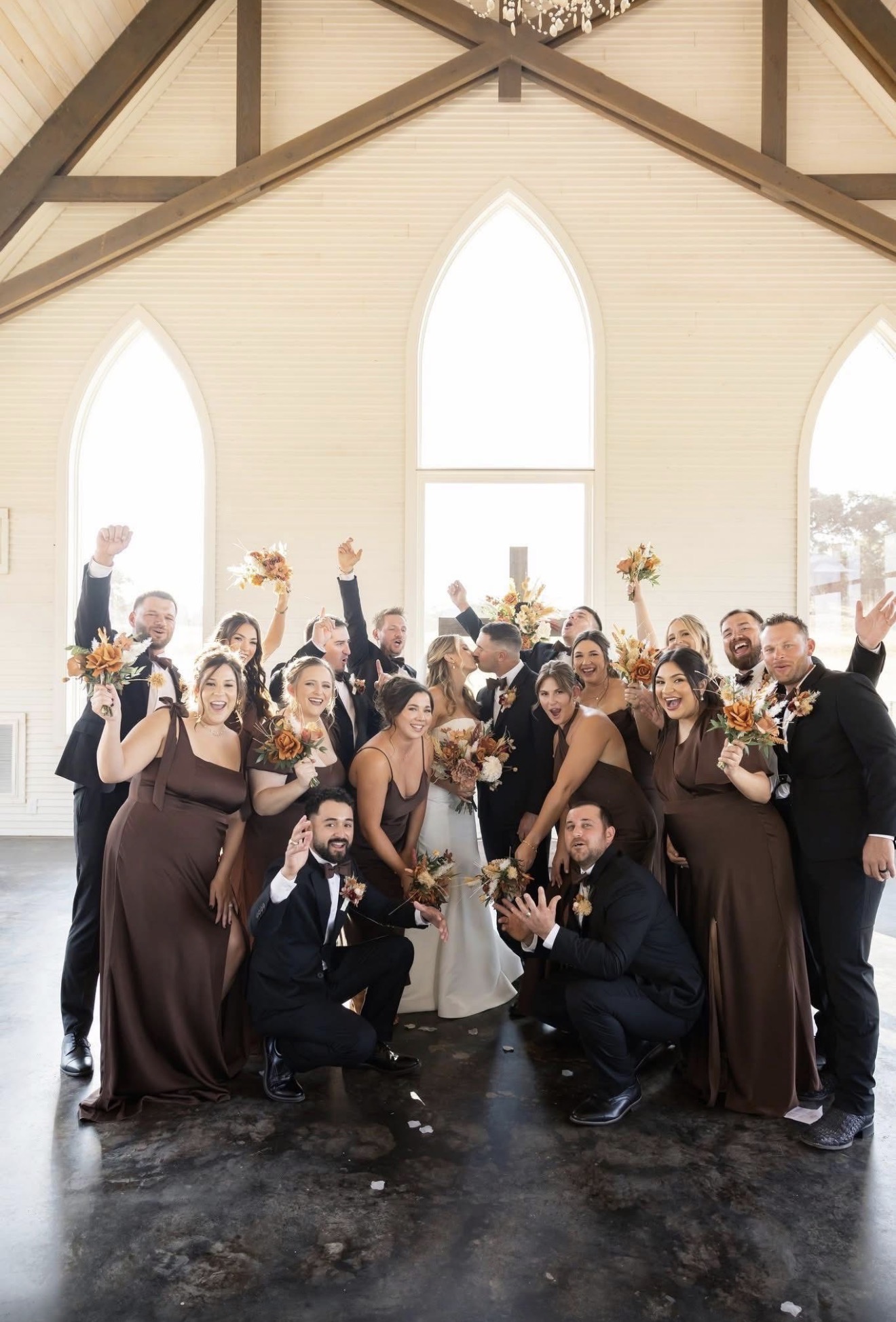 Group of people in formal attire inside a church with large arched windows, celebrating with bouquets and smiles.