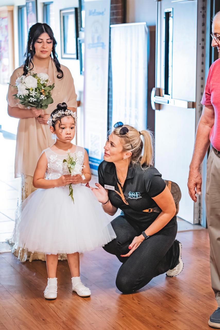 Young girl in a white dress holding a flower, woman crouching and talking to her, woman in beige dress holding flowers, person in pink shirt nearby, indoor setting