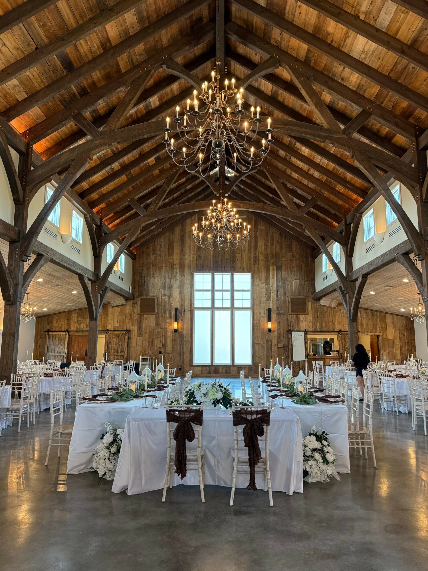 Elegant event space with high wooden ceiling, chandeliers, large window, and round tables with white tablecloths and floral centerpieces.