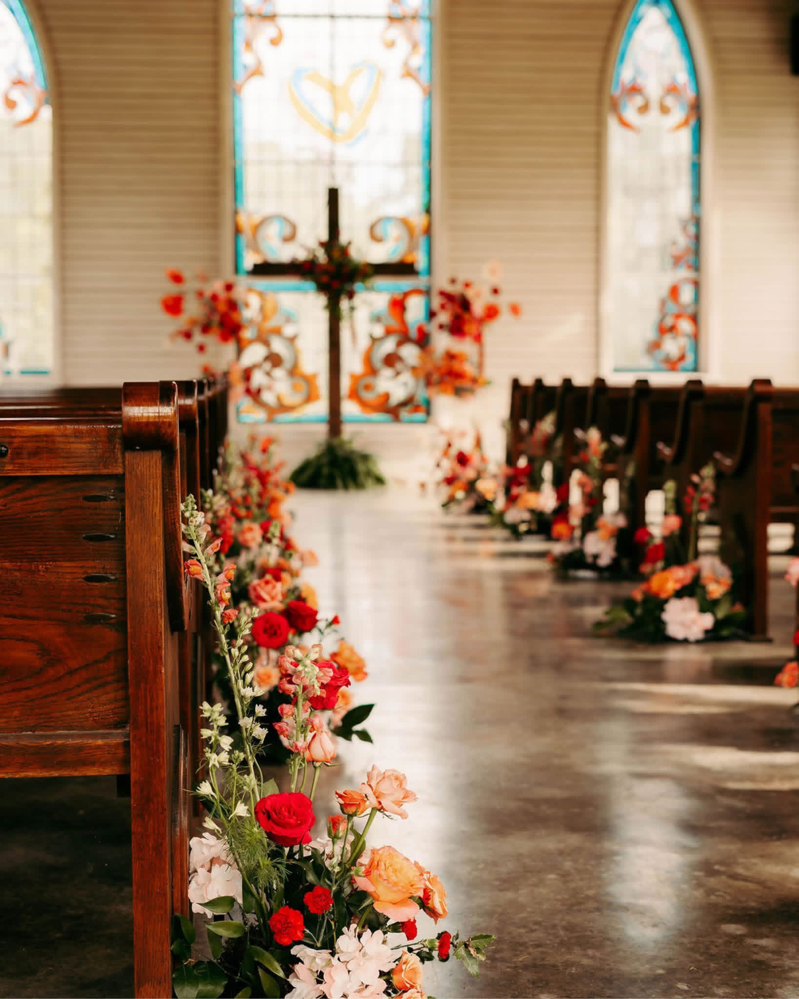 Interior of a church aisle decorated with flowers, leading to a cross and stained glass windows at the altar.
