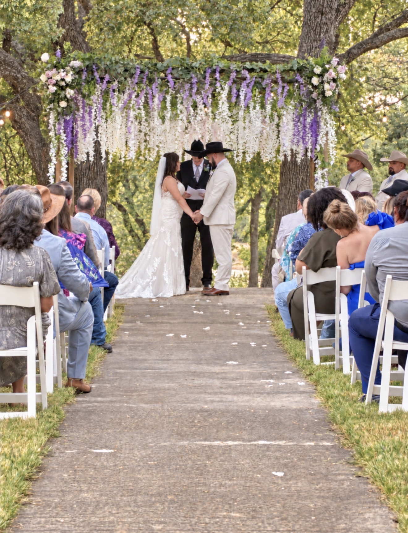 Couple holding hands at outdoor wedding ceremony under floral arch, guests seated on both sides, trees in background.