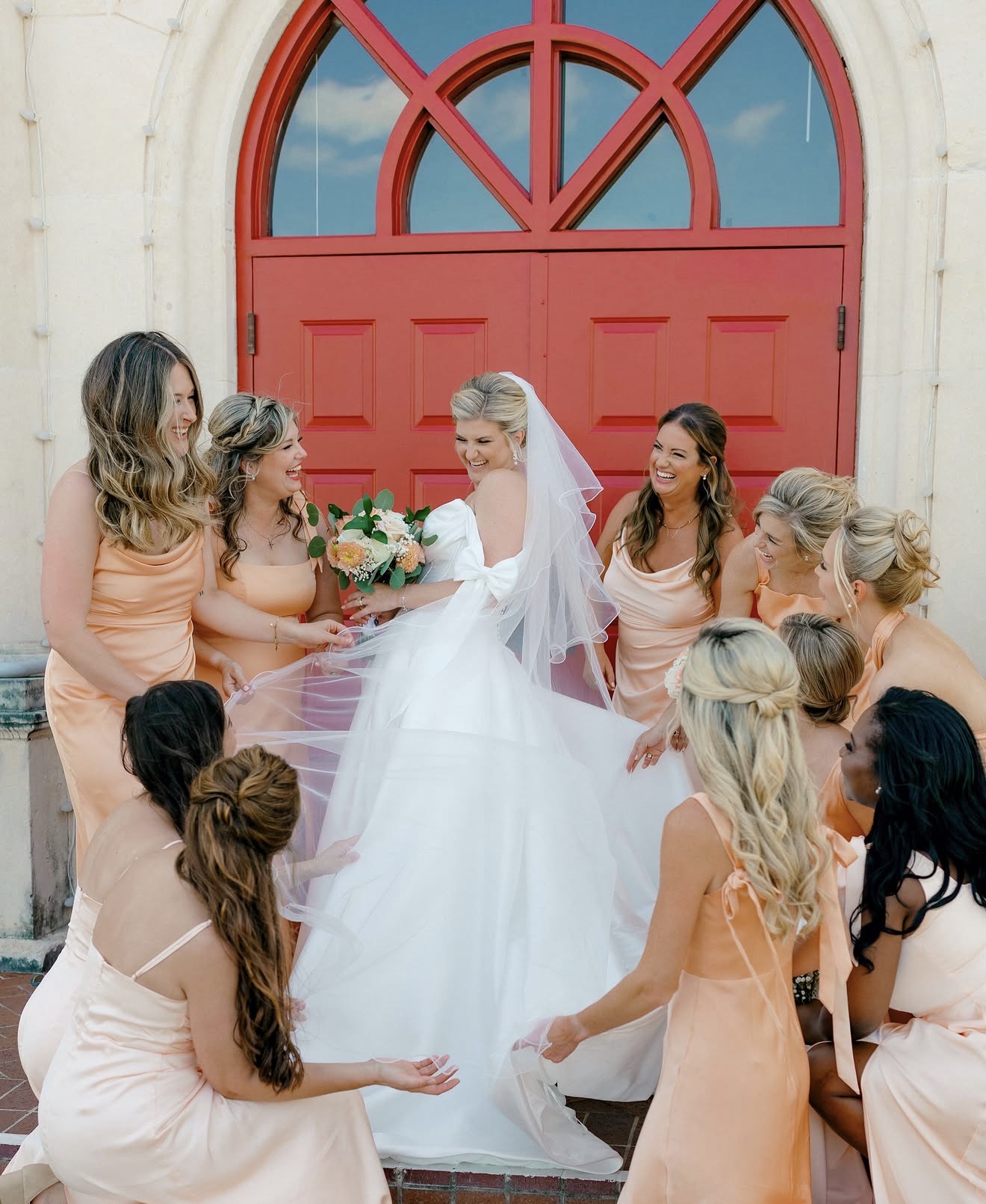 Bride in white dress surrounded by bridesmaids in peach dresses outside a building with a red arched door.