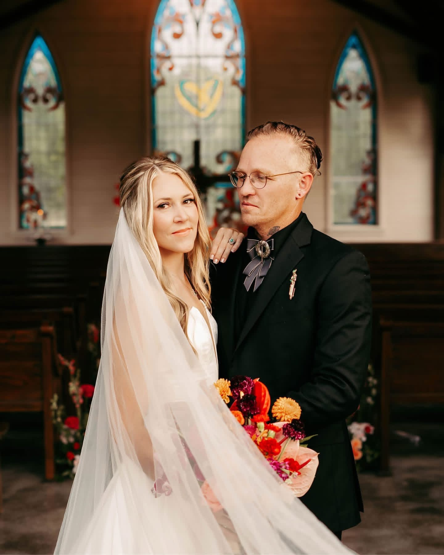 Bride and groom standing inside a church, bride holding a bouquet, stained glass windows in the background.