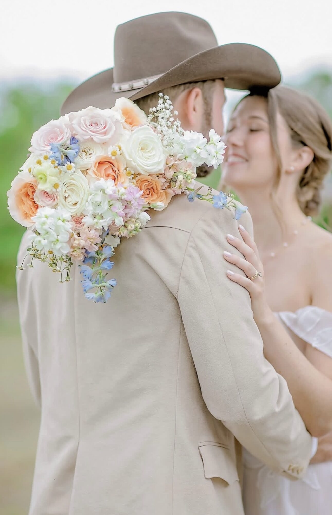 A couple faces each other outdoors, the man wearing a hat and beige suit, the woman smiling with her hand on his shoulder.