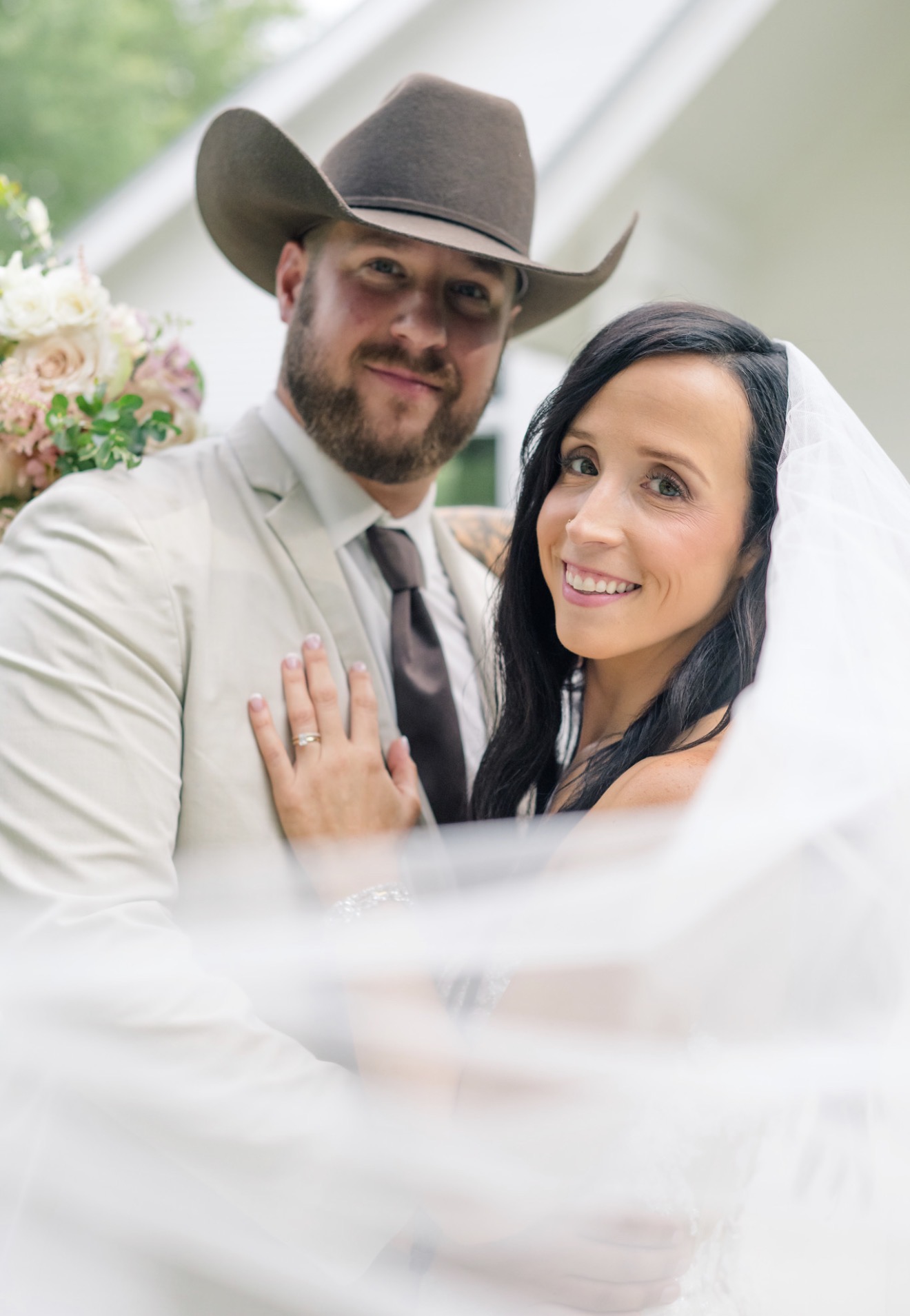 A smiling couple, the man wearing a cowboy hat and suit, the woman with dark hair, close together outdoors.