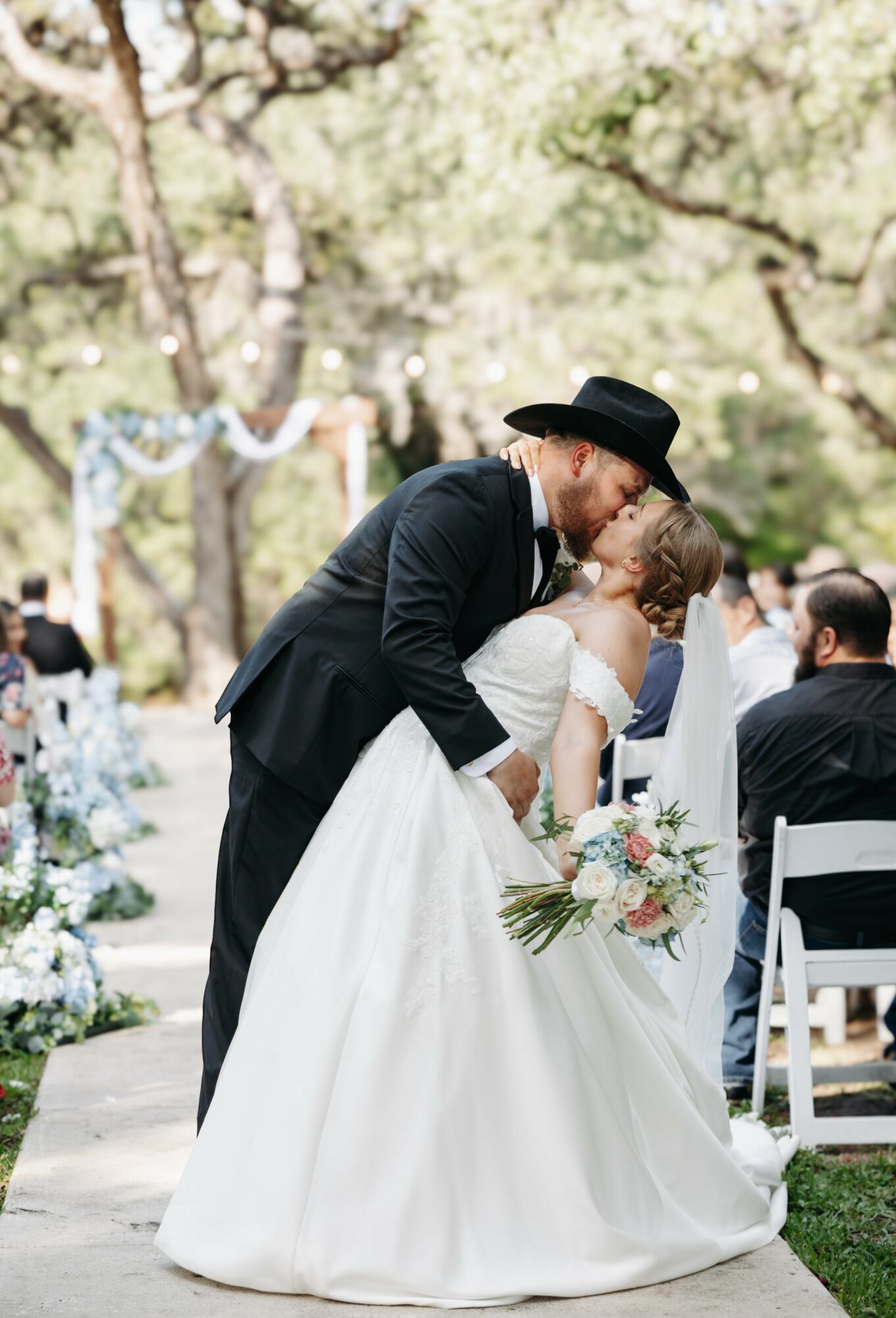 A couple shares a kiss at an outdoor wedding ceremony, surrounded by seated guests and decorated trees.