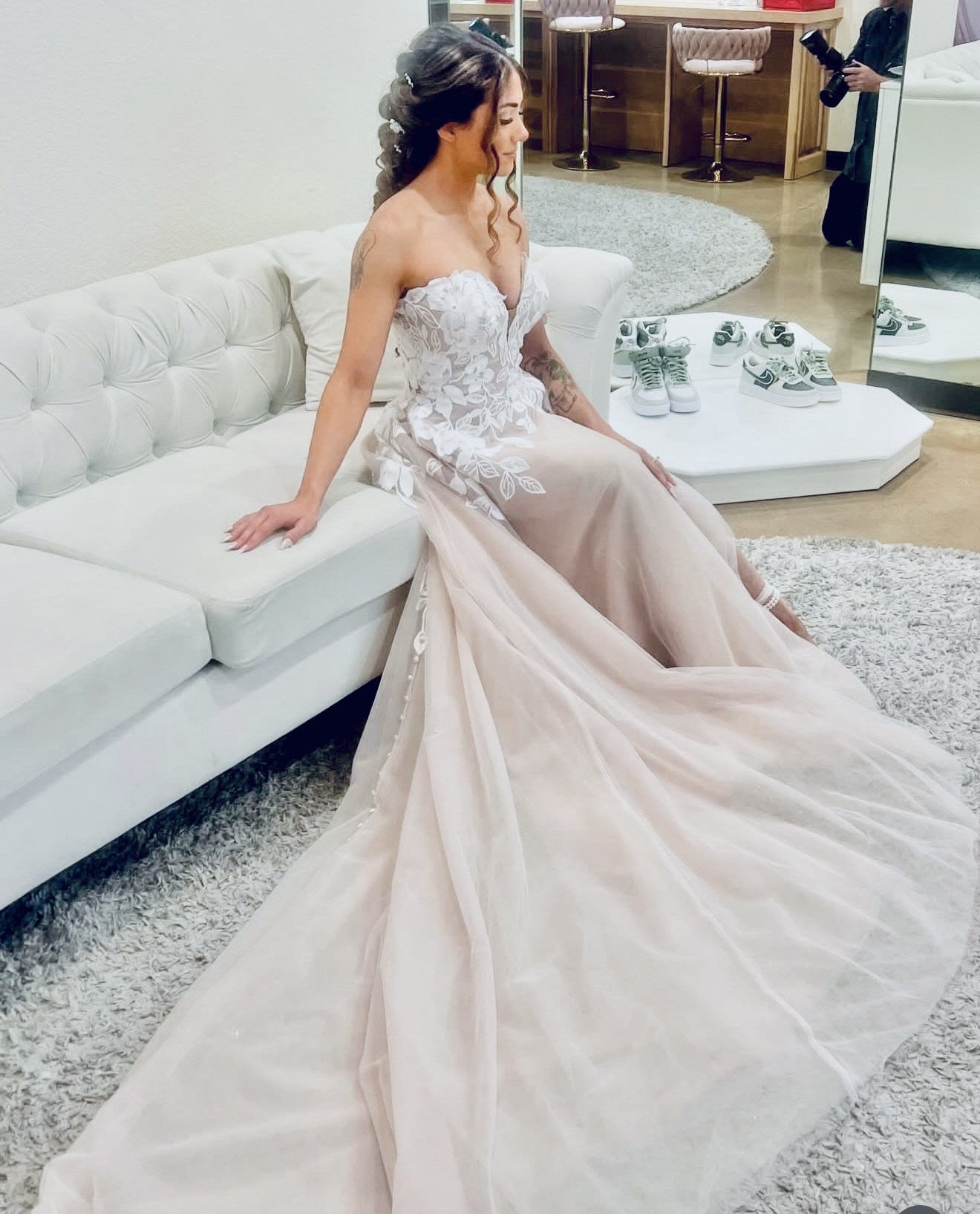 Woman in a wedding dress sitting on a white sofa, with a long train flowing on the floor, in a modern room.