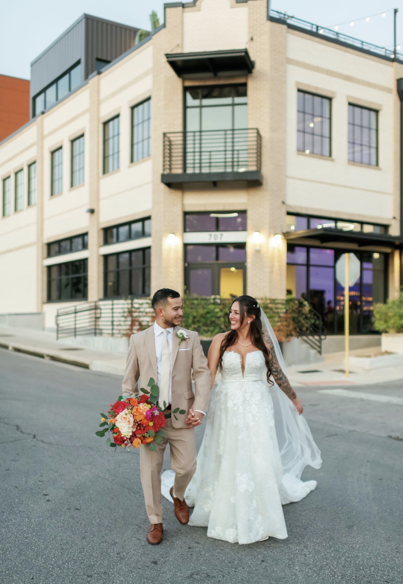 A bride and groom walk together on a city street, holding hands, with a modern building in the background.