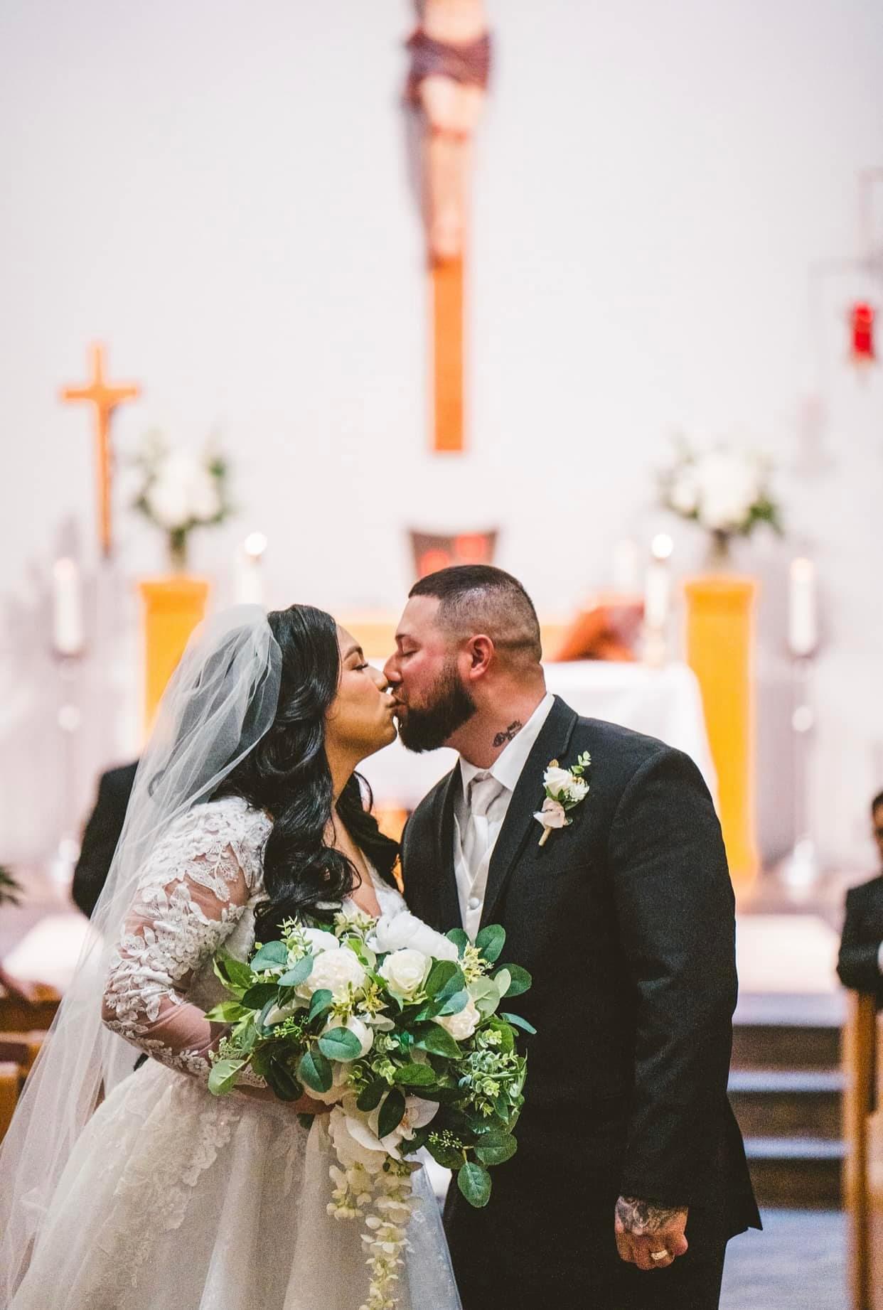Bride and groom kiss inside a church, bride holding a bouquet, altar with flowers and cross in background.