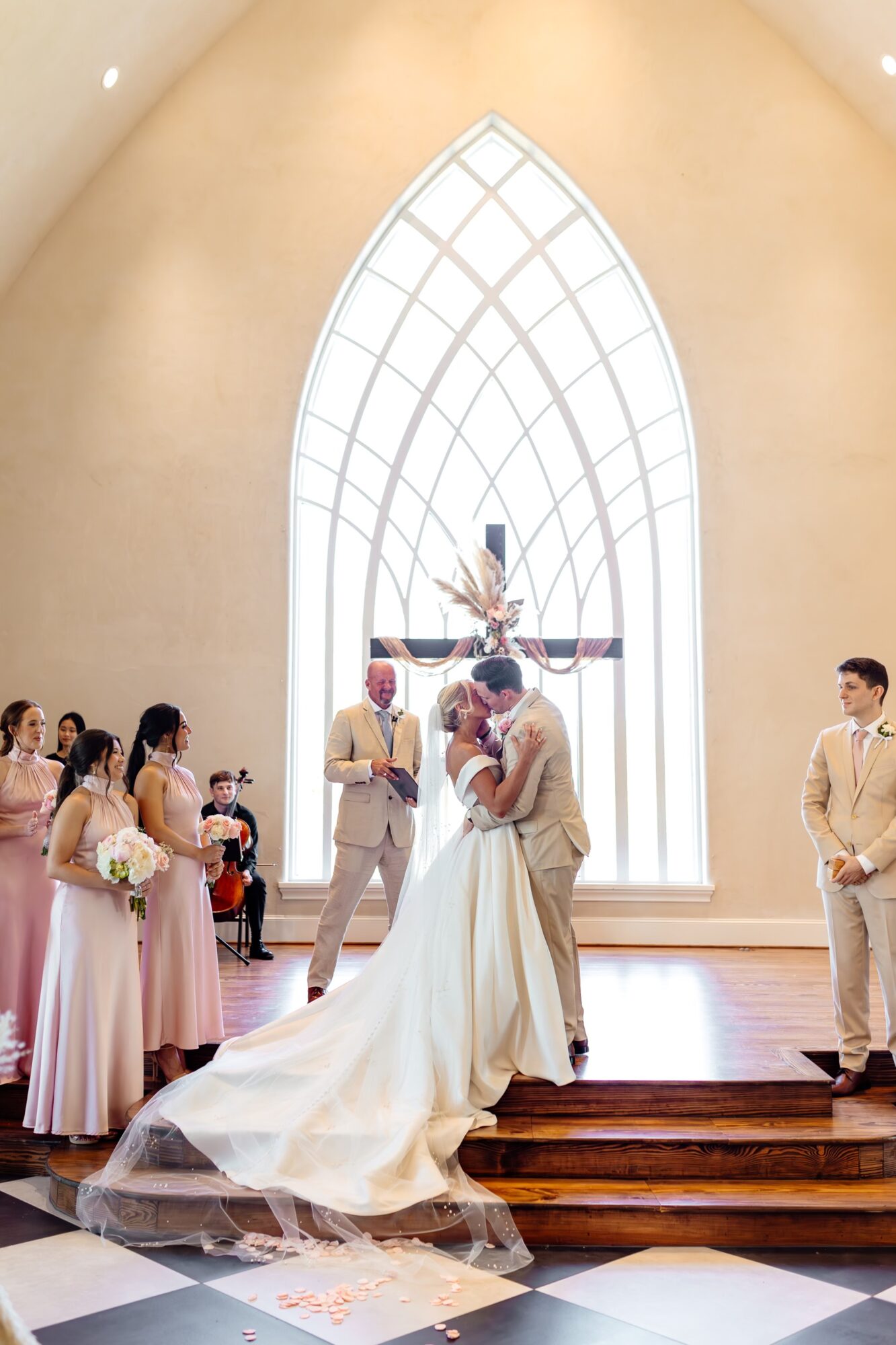 A wedding ceremony inside a church with a large stained glass window, bride and groom kissing, surrounded by bridesmaids and groomsmen.