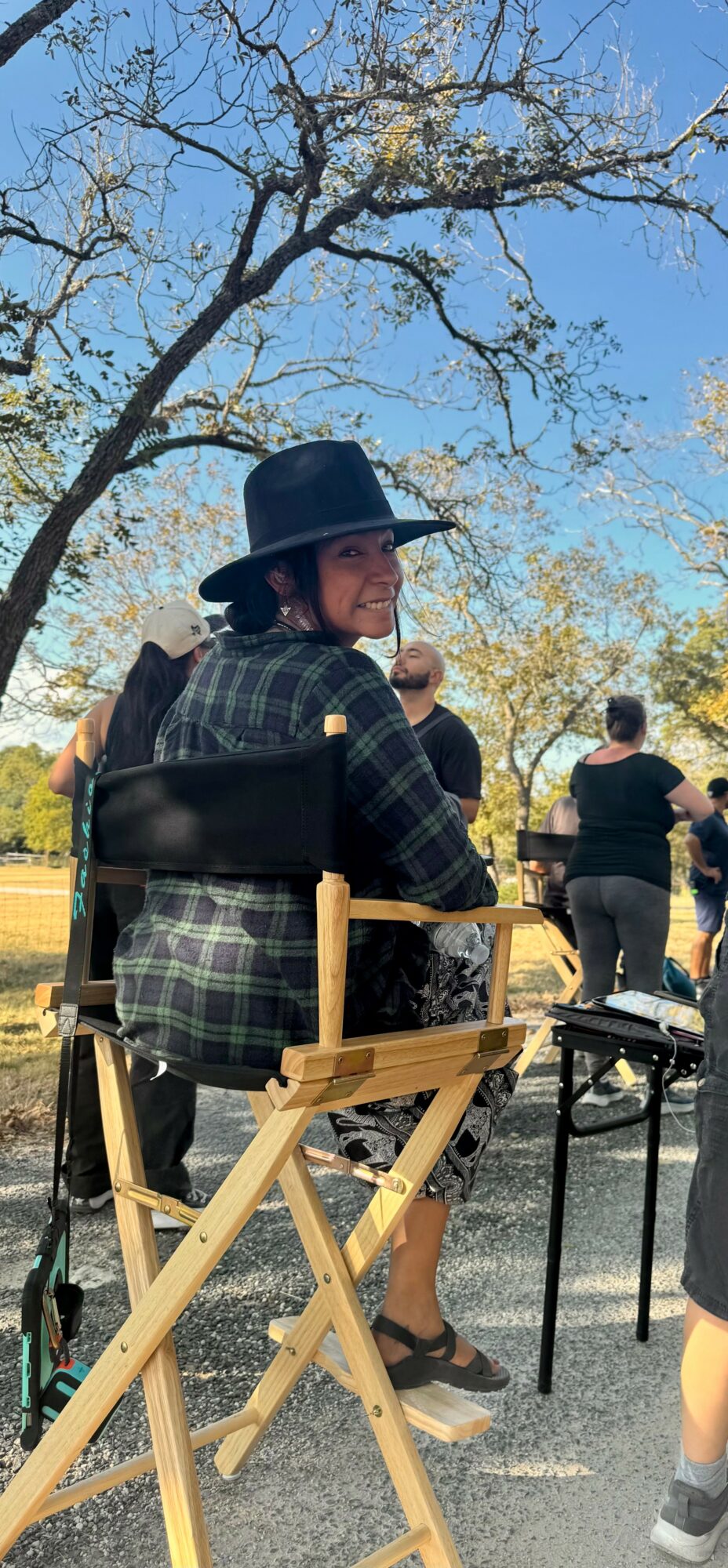 Person sitting on a wooden chair outdoors, wearing a black hat and plaid shirt, with trees and people in the background.