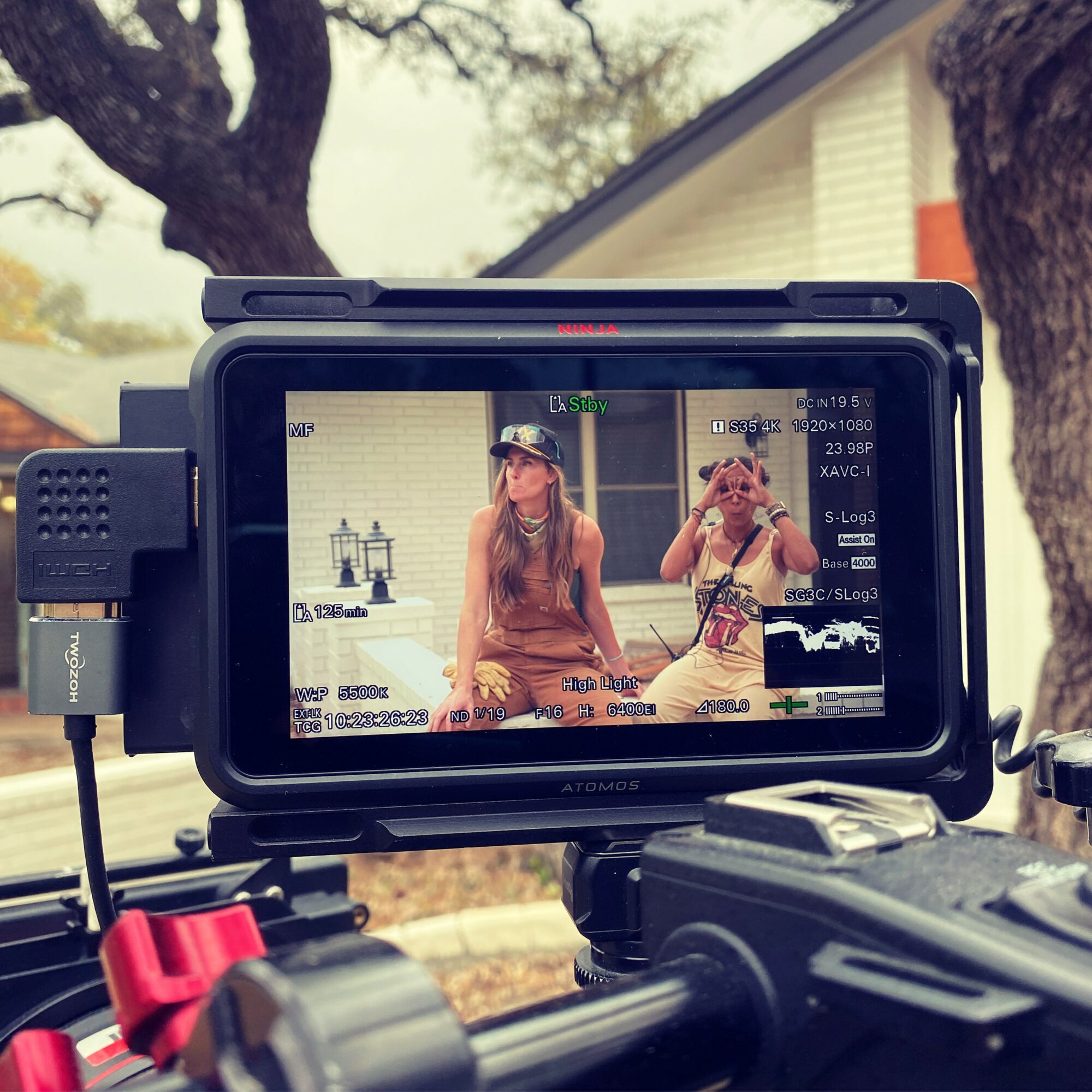 Two women sitting outdoors, one taking a photo with a camera, trees and a building in background.