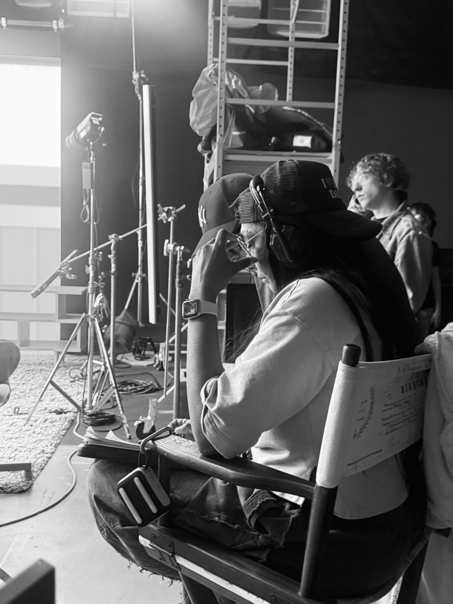 Person sitting on a chair with equipment and a backpack nearby, in a studio or stage setting, with a child and equipment in background.