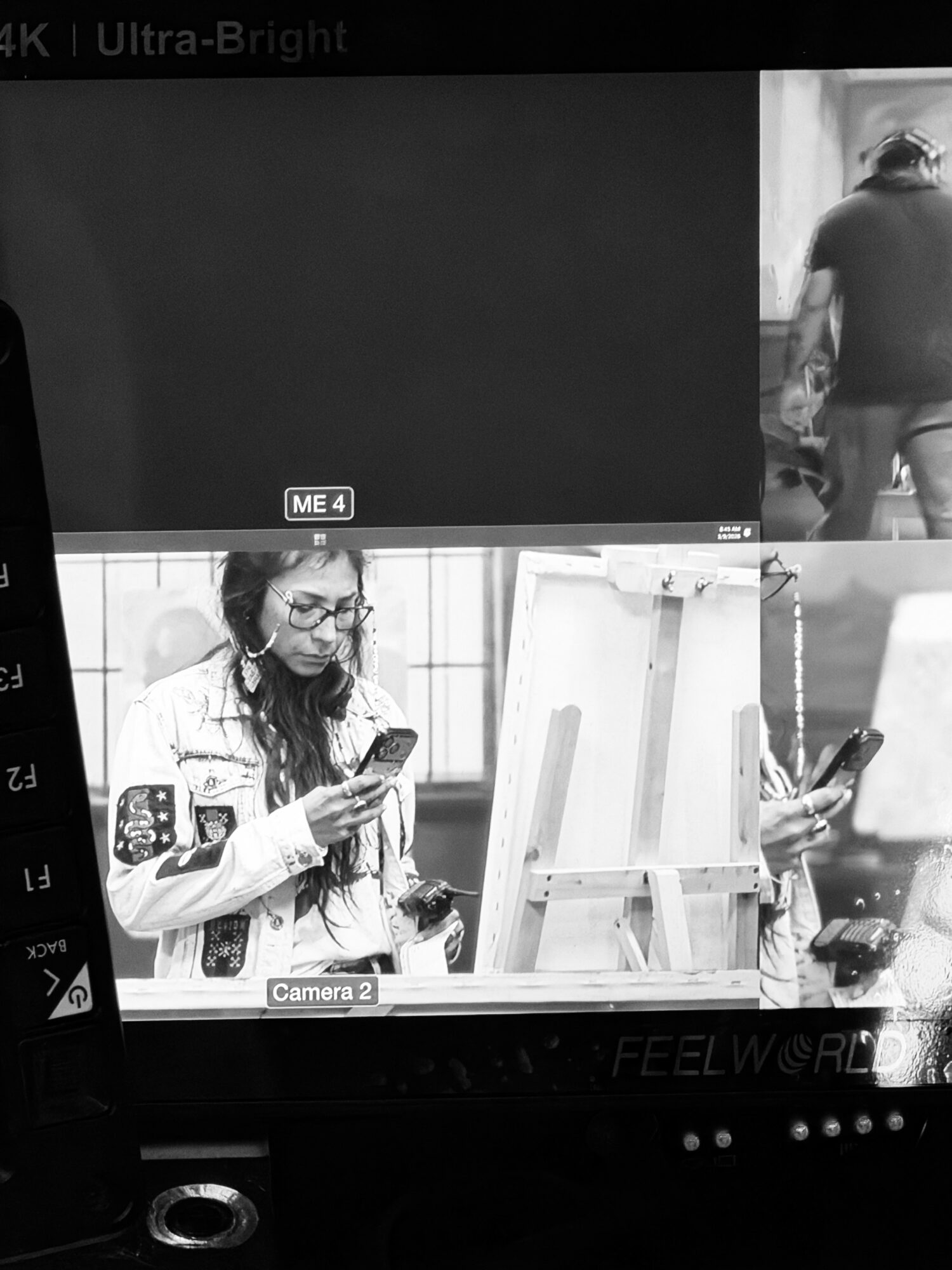 Woman with glasses looking at her phone, standing near an easel, in a bright indoor setting.