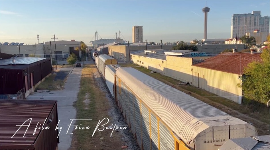 Train cars on tracks with city buildings and a tower in the background under a clear sky.