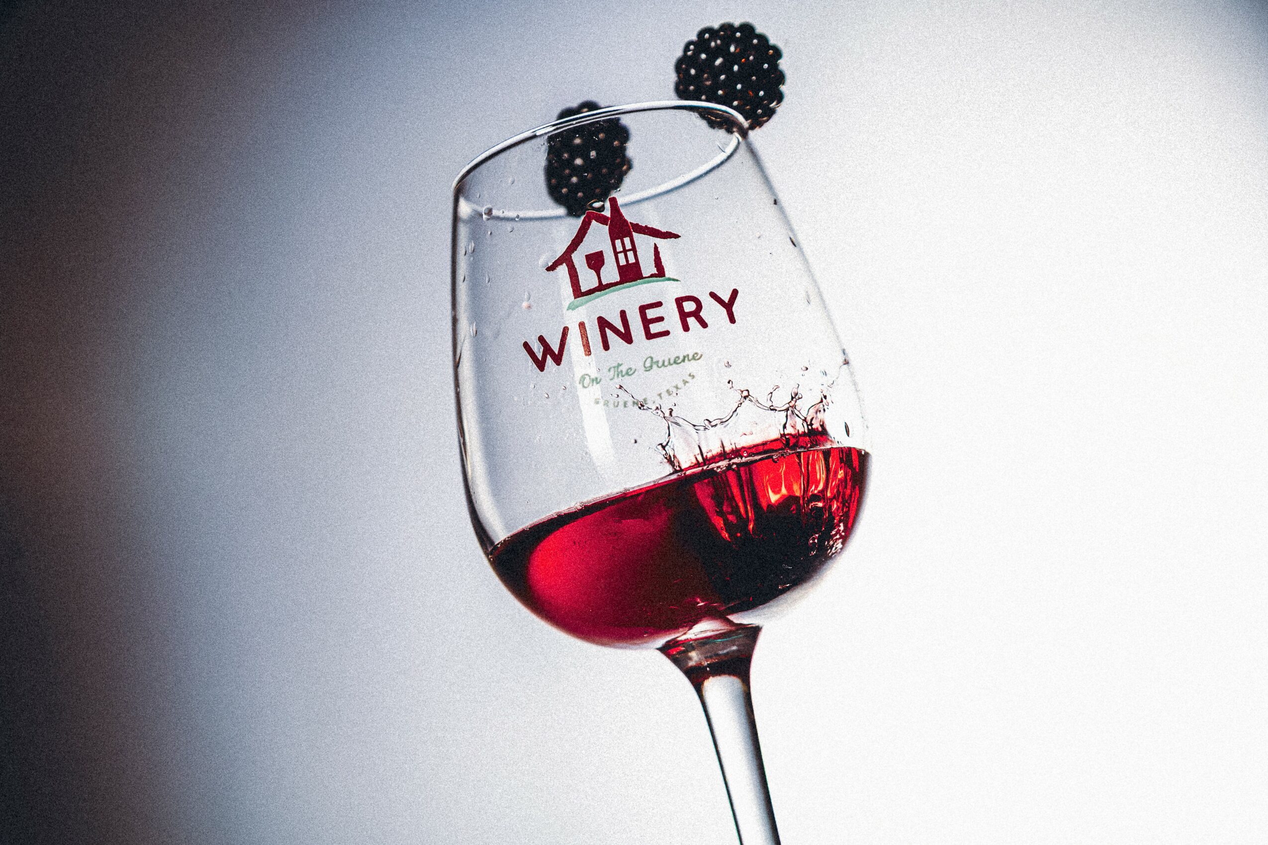 A wine glass with red wine, decorated with blackberries and a winery logo, against a plain background.
