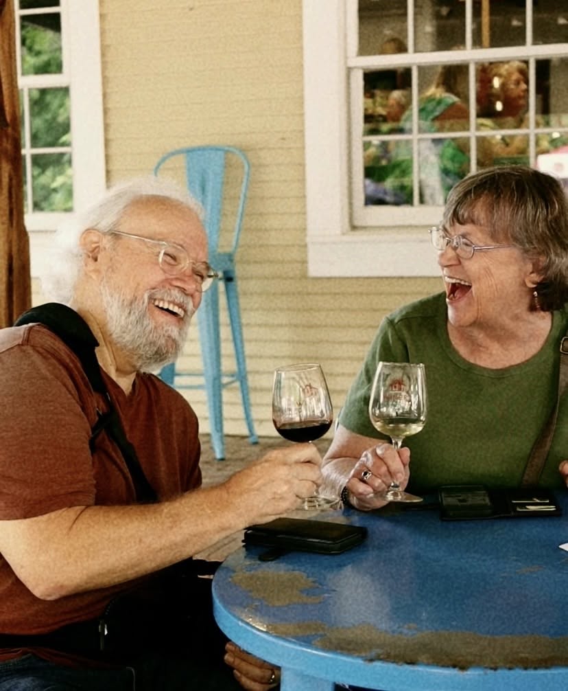 Two older adults, a man and a woman, sit at a blue table, smiling and holding glasses of red wine.