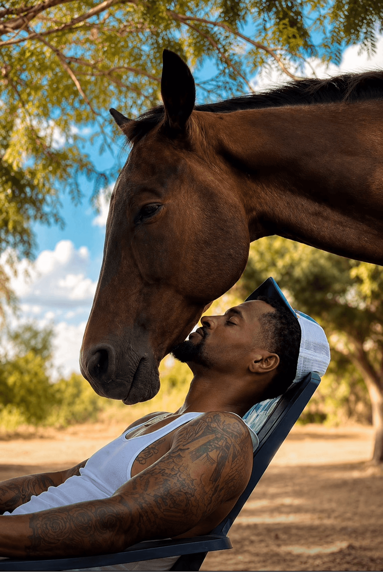 Man with tattoos and a cap leans back in a chair outdoors, kissing a large brown horse's nose under a tree.