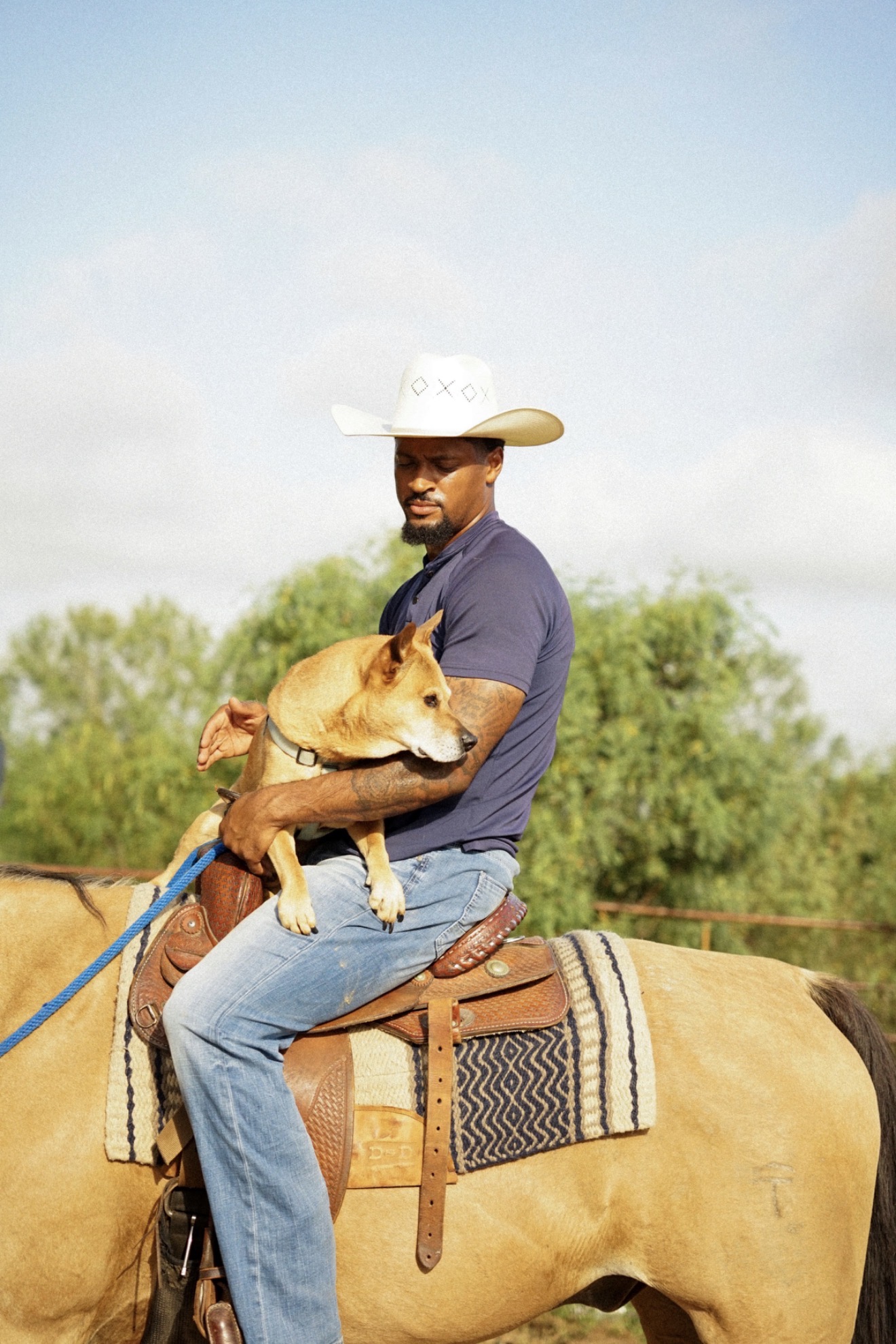 Man wearing a cowboy hat sitting on a horse, holding a dog, outdoors with trees and sky in background.
