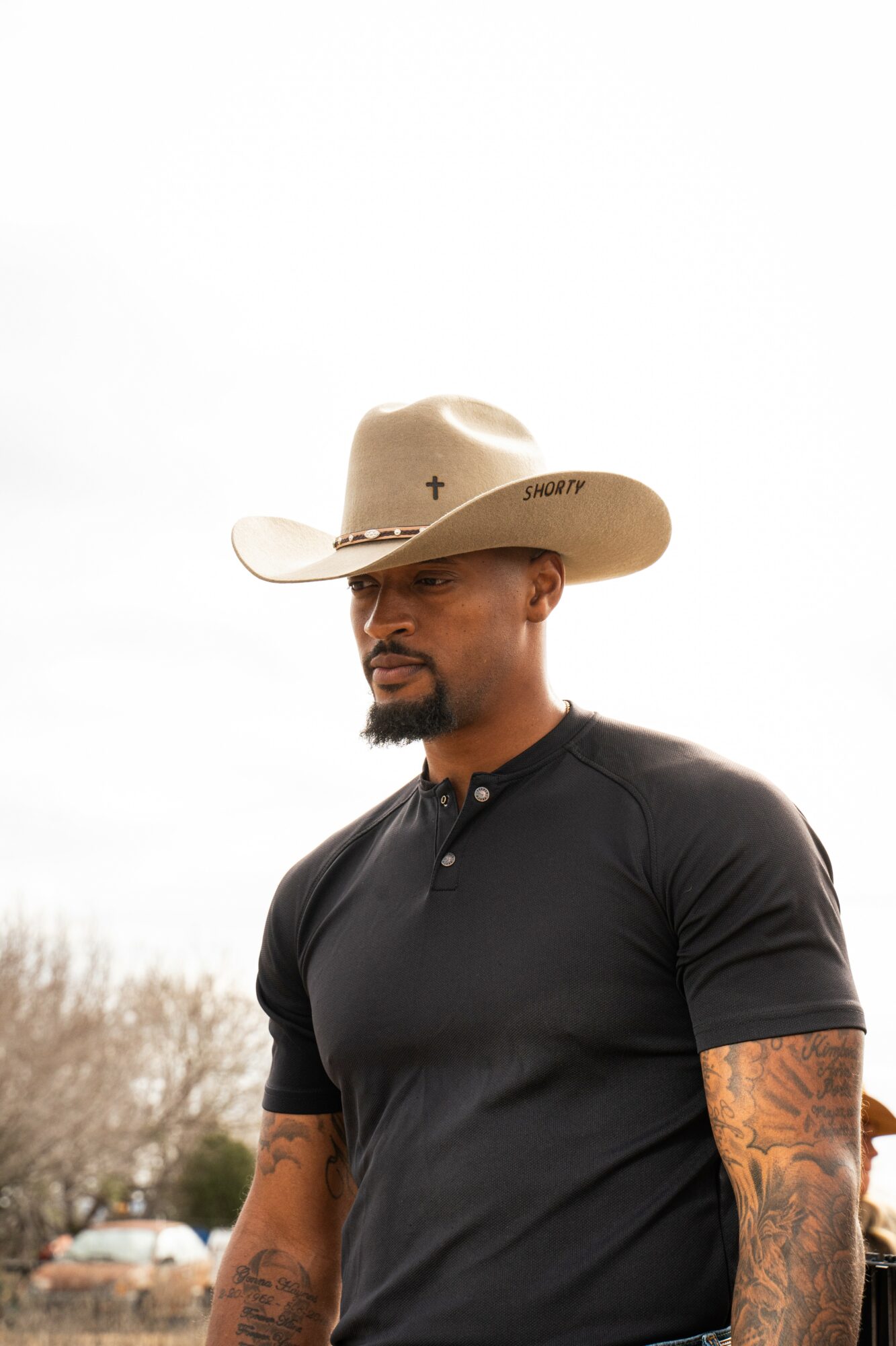 Man with a beard and tattoos wearing a beige cowboy hat and black shirt, standing outdoors against a cloudy sky.