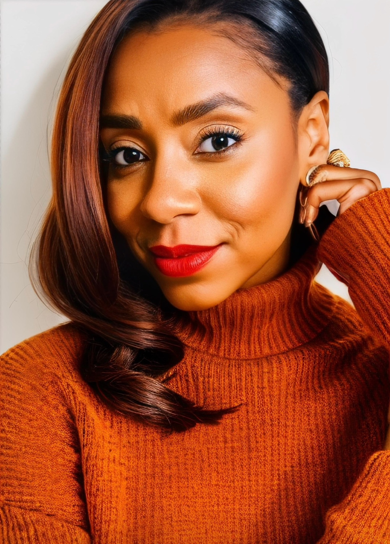 Close-up of a woman with smooth hair, wearing a rust-colored turtleneck, smiling with red lipstick, touching her face.