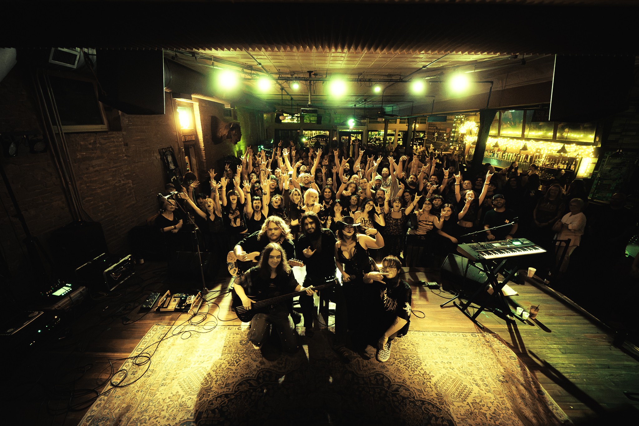 Group of people on stage in a dimly lit venue, with musical equipment and a crowd in the background.