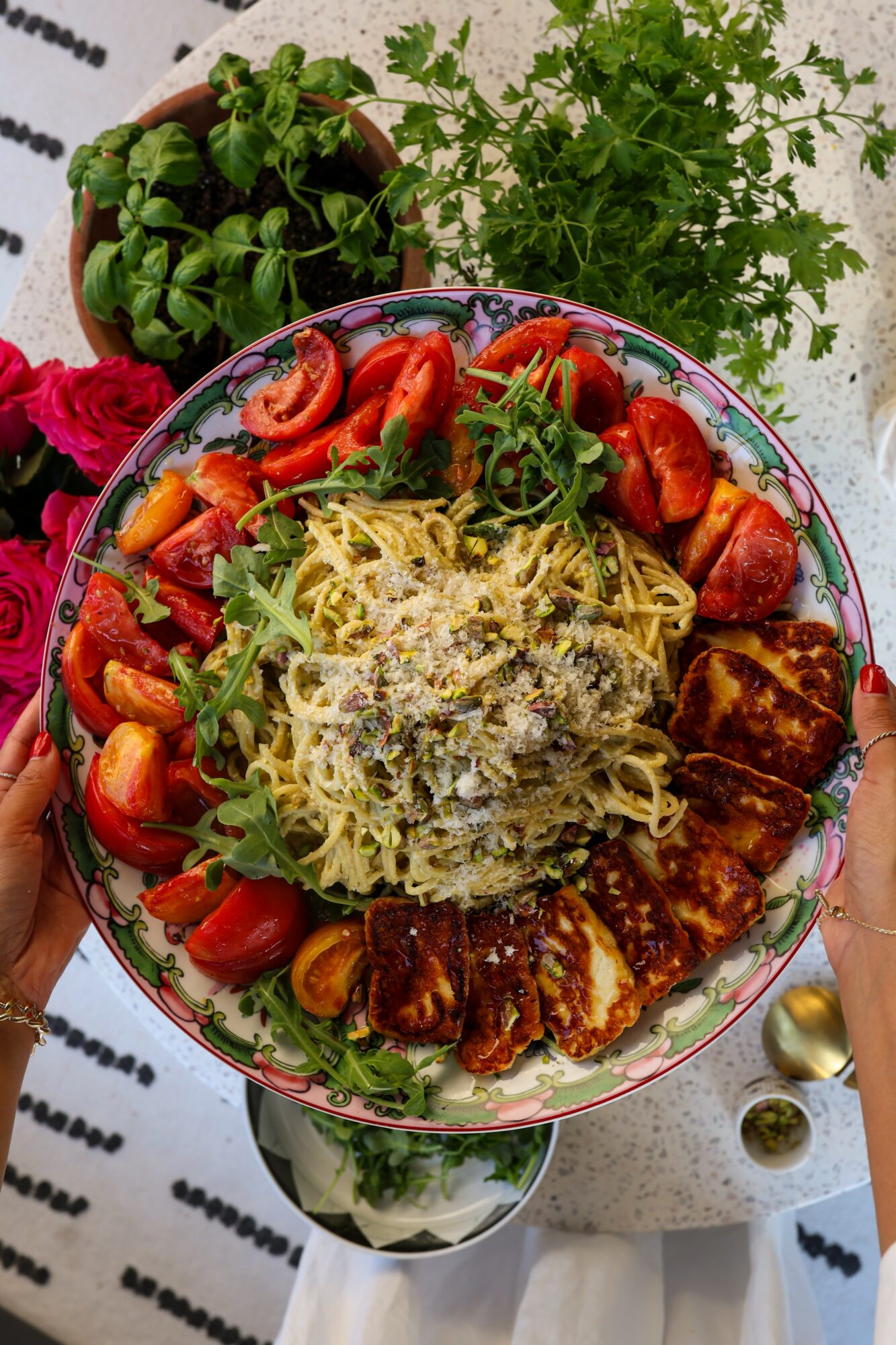 Plate with pasta, strawberries, grilled chicken, and greens, held over a table with potted plants in the background.