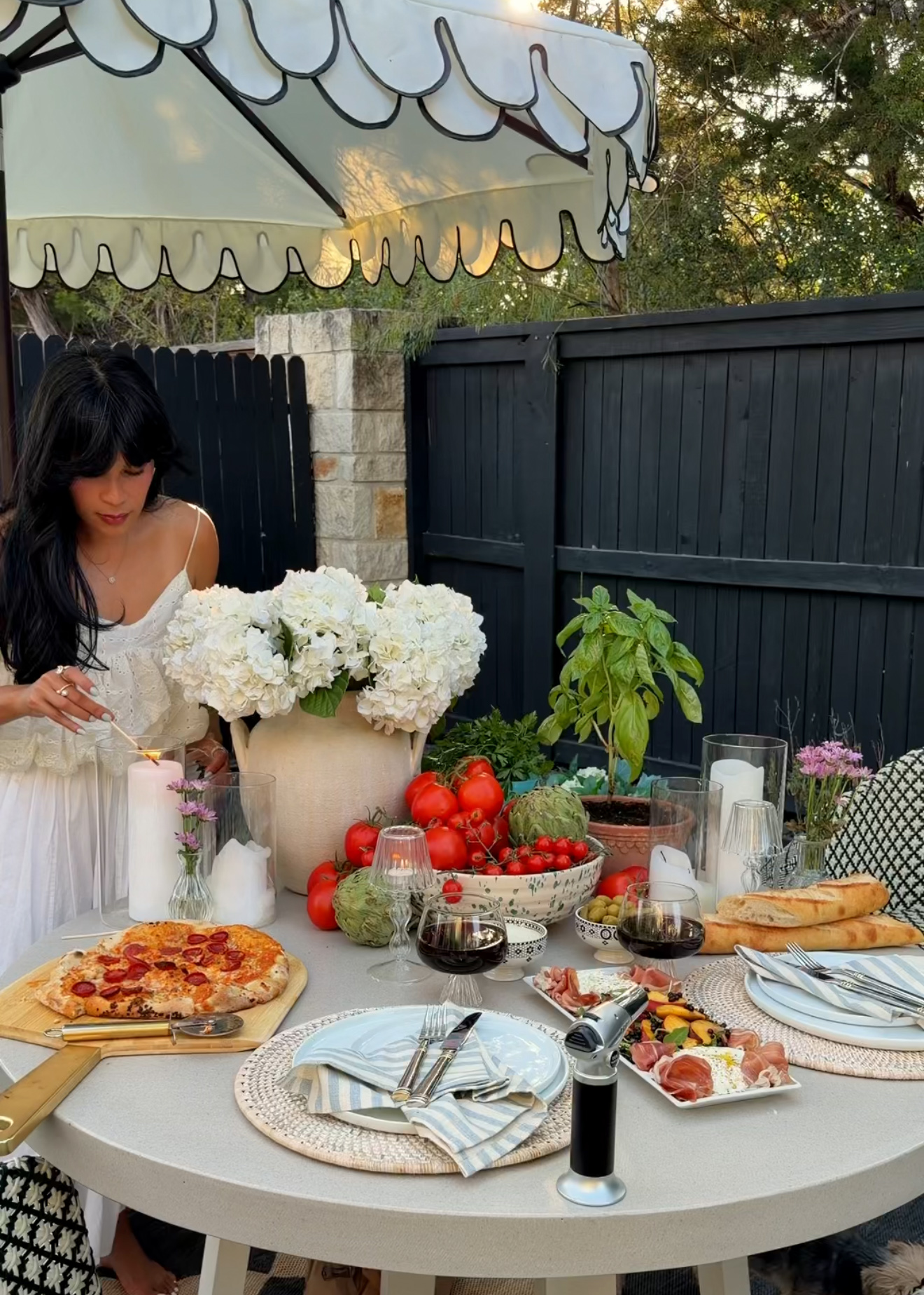 Outdoor table set with pizza, flowers, fruits, candles, and utensils, surrounded by a black fence and trees.